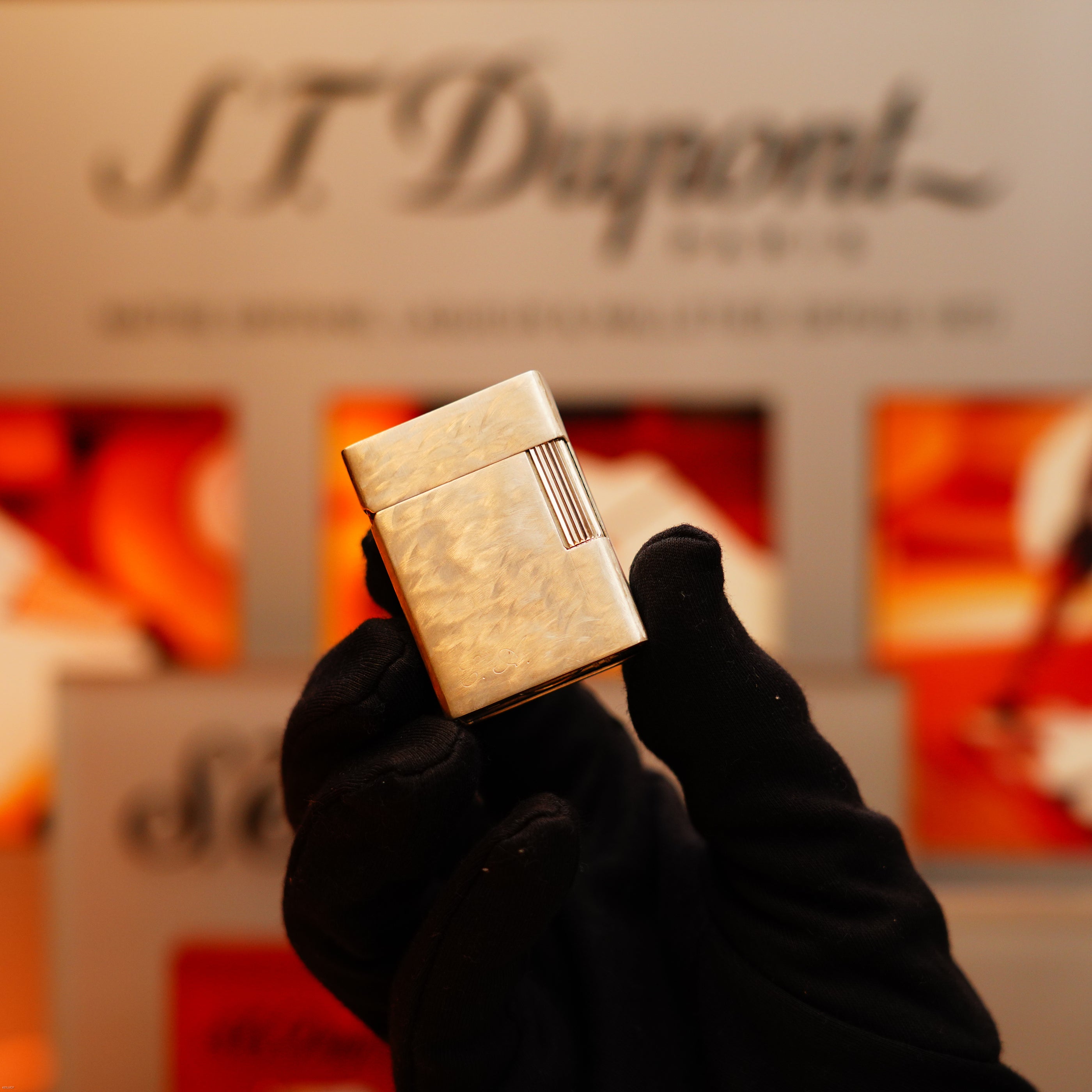 A gloved hand holds a Vintage 1970 de St Dupont Two Tone Solid 925 Sterling Silver Guilloché Pattern Lighter by S.T. Dupont in front of a blurred S.T. Dupont display.