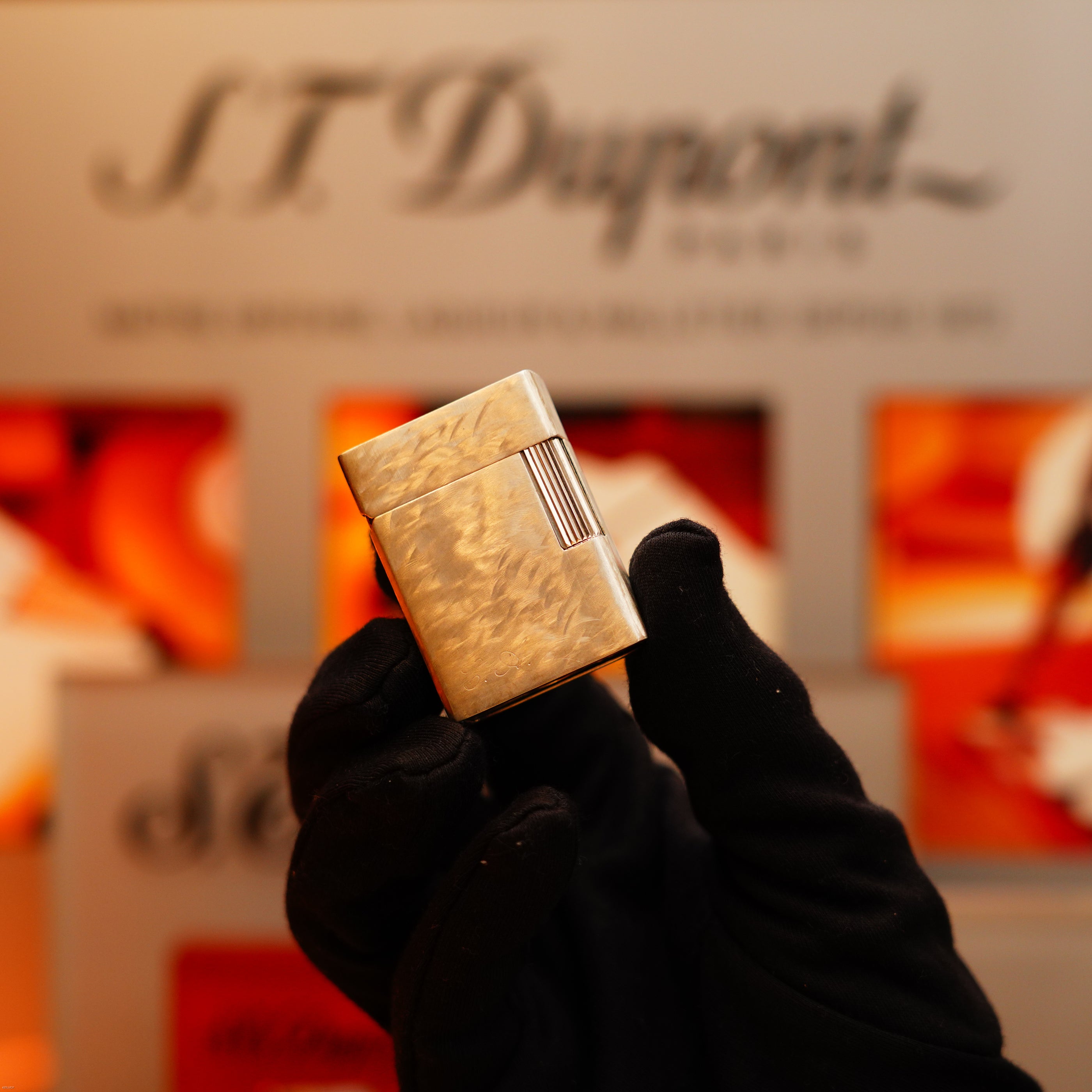 A gloved hand holds a Vintage 1970 de S.T. Dupont Two Tone Solid 925 Sterling Silver Guilloché Pattern Lighter in front of an S.T. Dupont display.