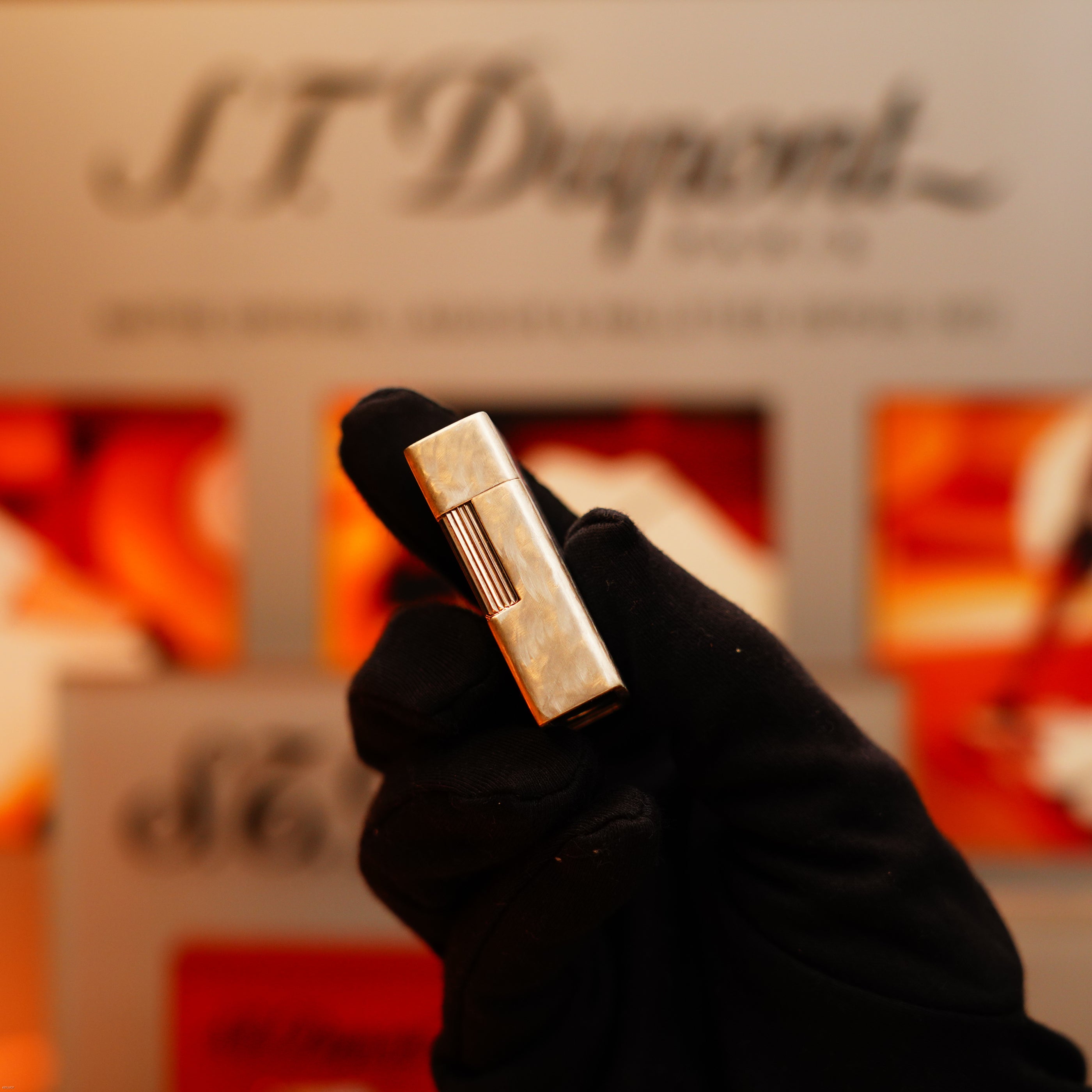 A gloved hand holds a Vintage 1970 de St Dupont Two Tone Solid 925 Sterling Silver Guilloché Pattern Lighter by S.T. Dupont, with a blurred S.T. Dupont display in the background.