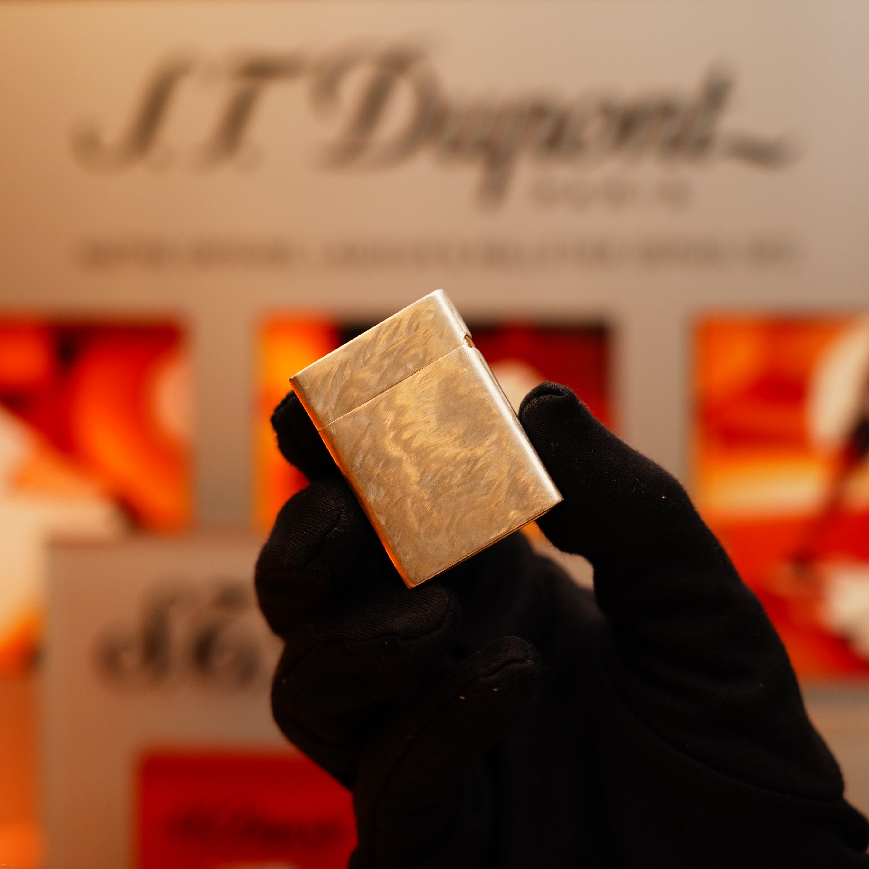 A gloved hand holds a Vintage 1970 de St Dupont Two Tone Solid 925 Sterling Silver Guilloché Pattern Lighter by S.T. Dupont in front of a blurred brand display.