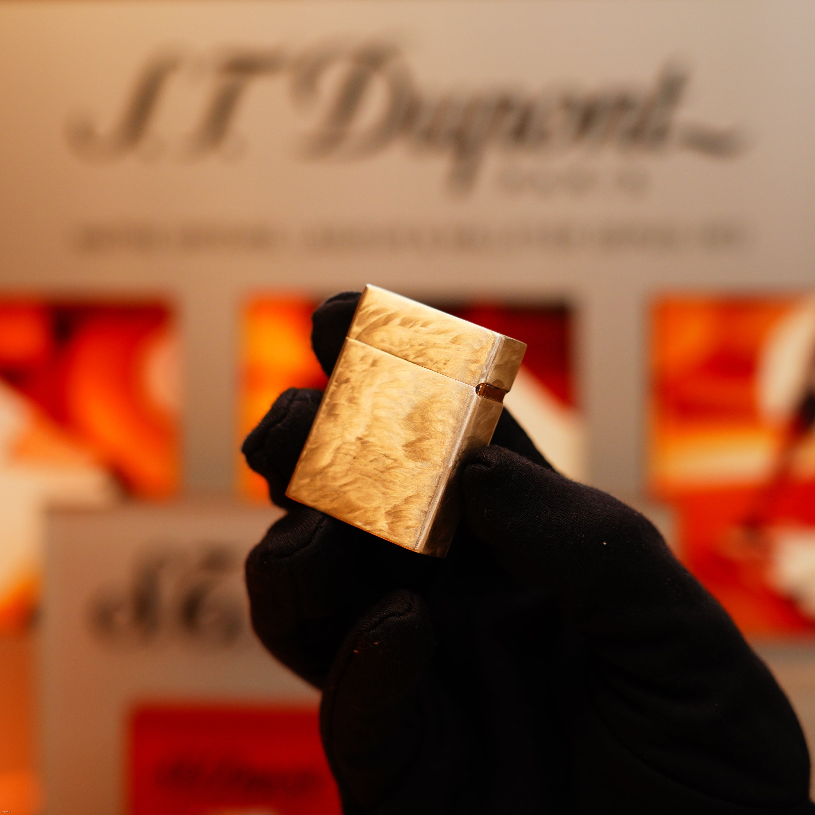 A gloved hand holds a Vintage 1970 S.T. Dupont Two Tone Solid 925 Sterling Silver Guilloché Pattern Lighter before a blurred S.T. Dupont sign.