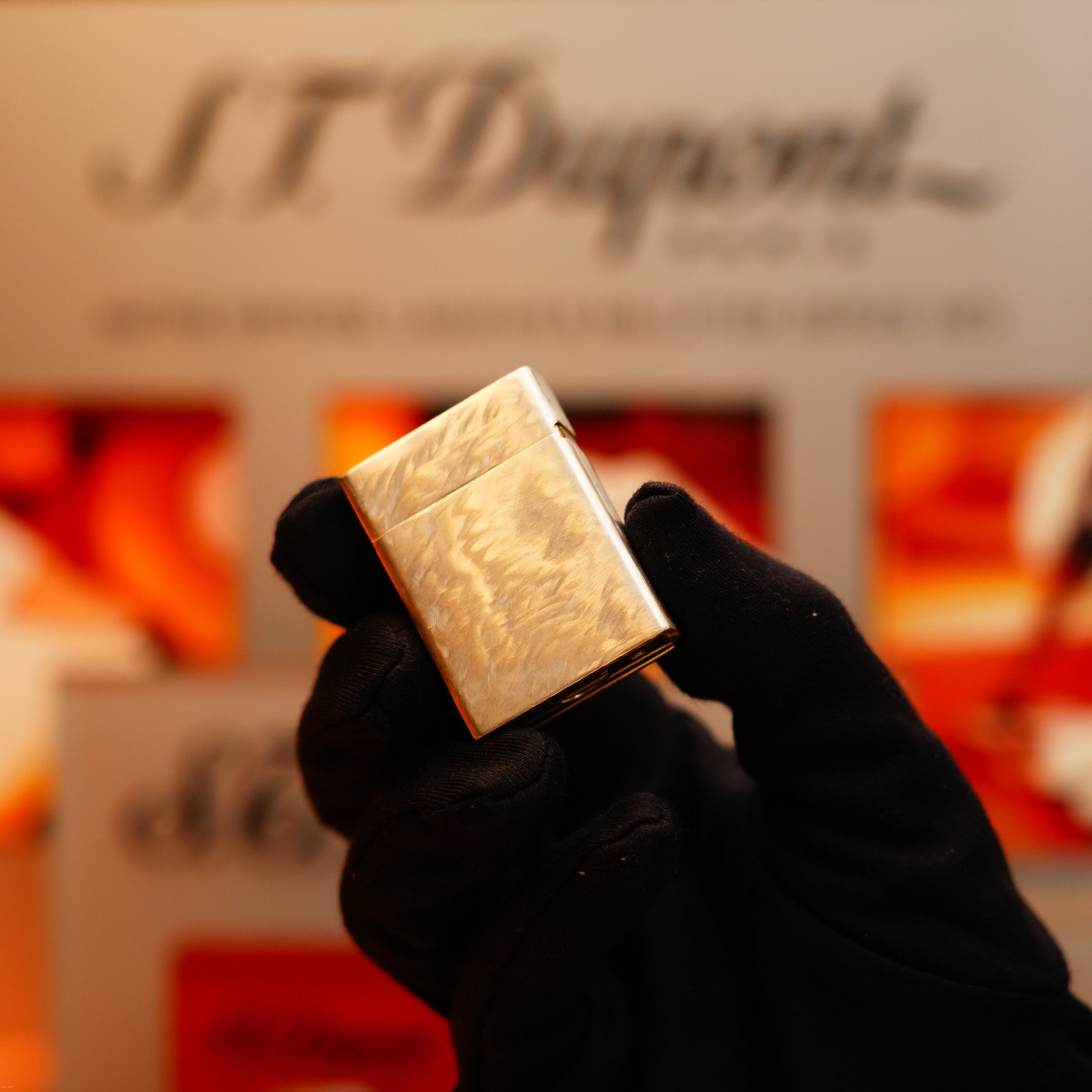 A gloved hand holds the Vintage 1970 de St Dupont Two Tone Solid 925 Sterling Silver Guilloché Pattern Lighter by S.T. Dupont in front of a blurred, orange-toned background with the S.T. Dupont logo visible.