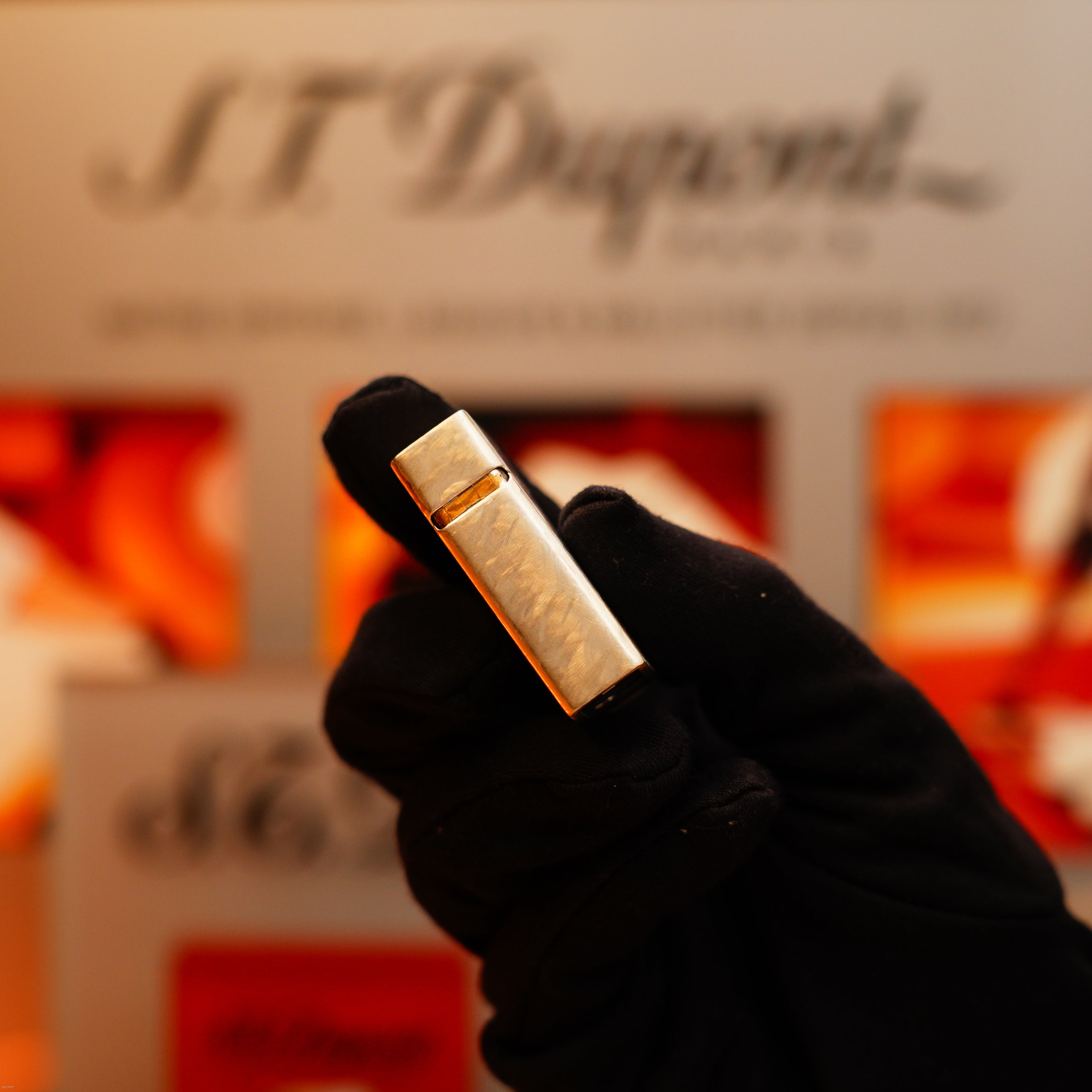 A gloved hand holds a Vintage 1970 de St Dupont Two Tone Solid 925 Sterling Silver Guilloché Pattern Lighter by S.T. Dupont, with a blurred S.T. Dupont display in the background.