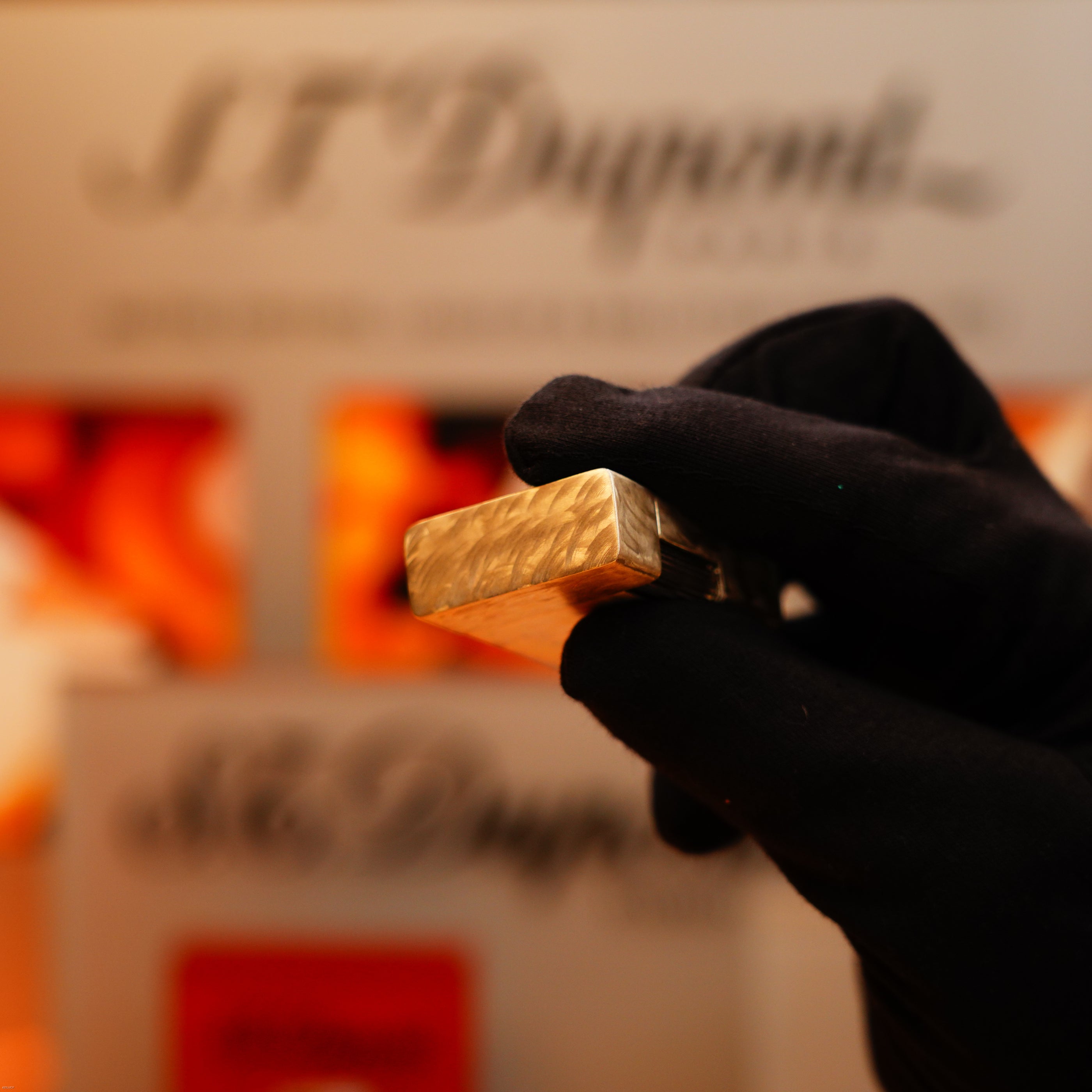 A gloved hand holds a Vintage 1970 de St Dupont Two Tone Solid 925 Sterling Silver Guilloché Pattern Lighter in front of a blurry S.T. Dupont sign.