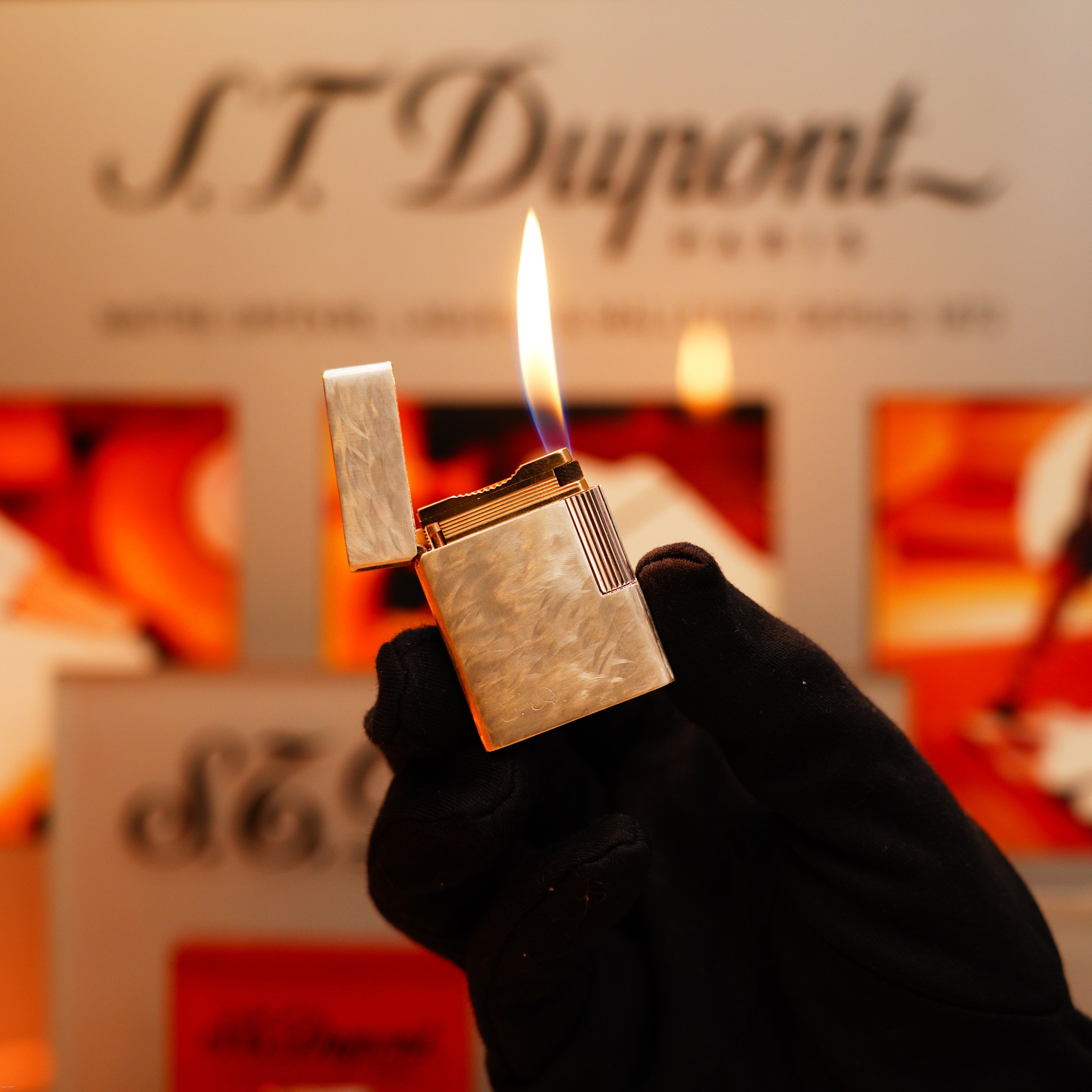 A gloved hand holds a lit S.T. Dupont Vintage 1970 Two Tone Solid 925 Sterling Silver Guilloché Pattern Lighter in front of a blurred S.T. Dupont logo background.