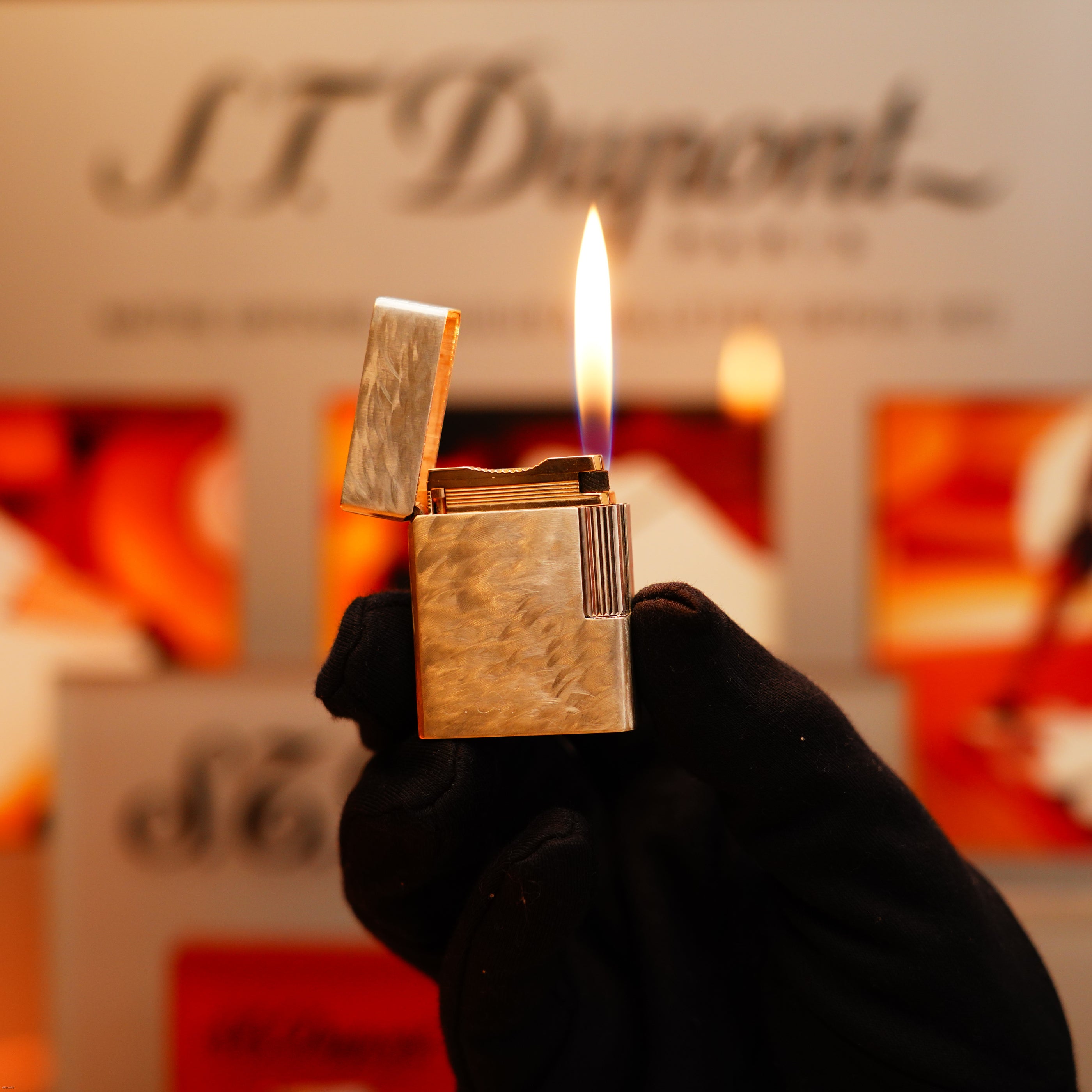 A gloved hand holds a lit Vintage 1970 de St Dupont Two Tone Solid 925 Sterling Silver Guilloché Pattern Lighter. In the blurred background, the S.T. Dupont logo and red-orange accents are visible.