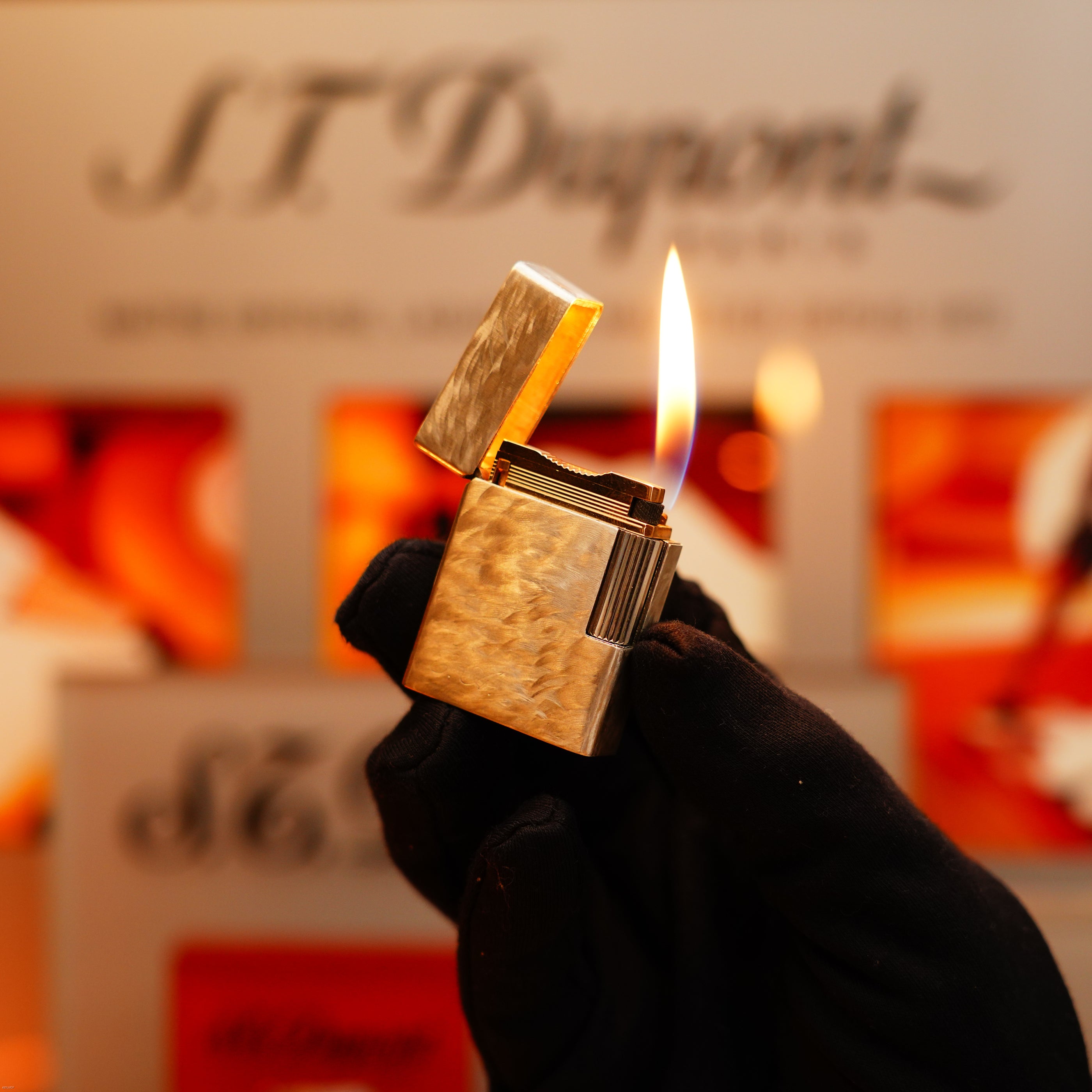 A gloved hand holds an open, lit Vintage 1970 de St Dupont Two Tone Solid 925 Sterling Silver Guilloché Pattern Lighter, with a visible flame; the blurred S.T. Dupont logo appears in the background.