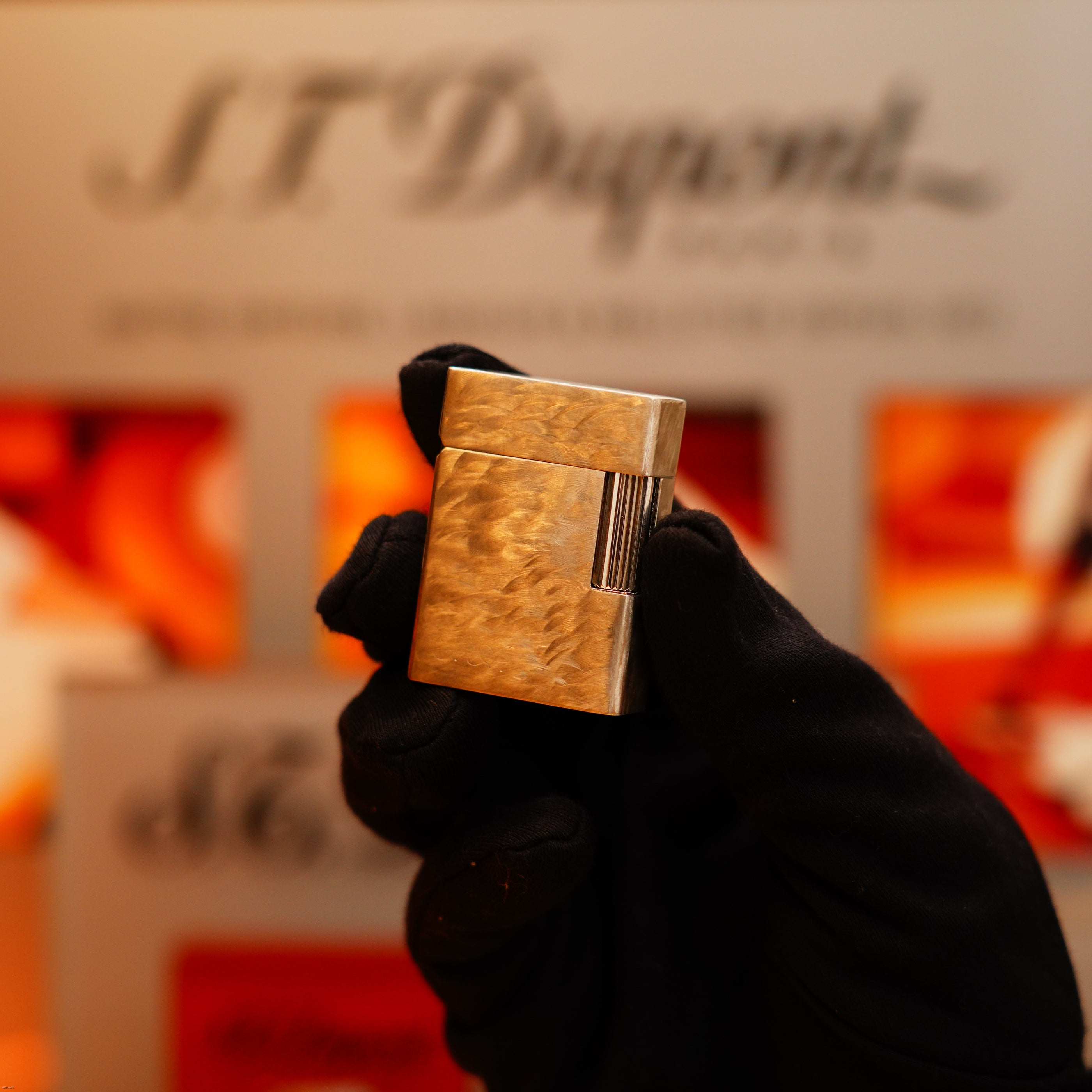 A hand in a black glove holds a Vintage 1970 de St Dupont Two Tone Solid 925 Sterling Silver Guilloché Pattern Lighter by S.T. Dupont, with a blurred branded display in the background.