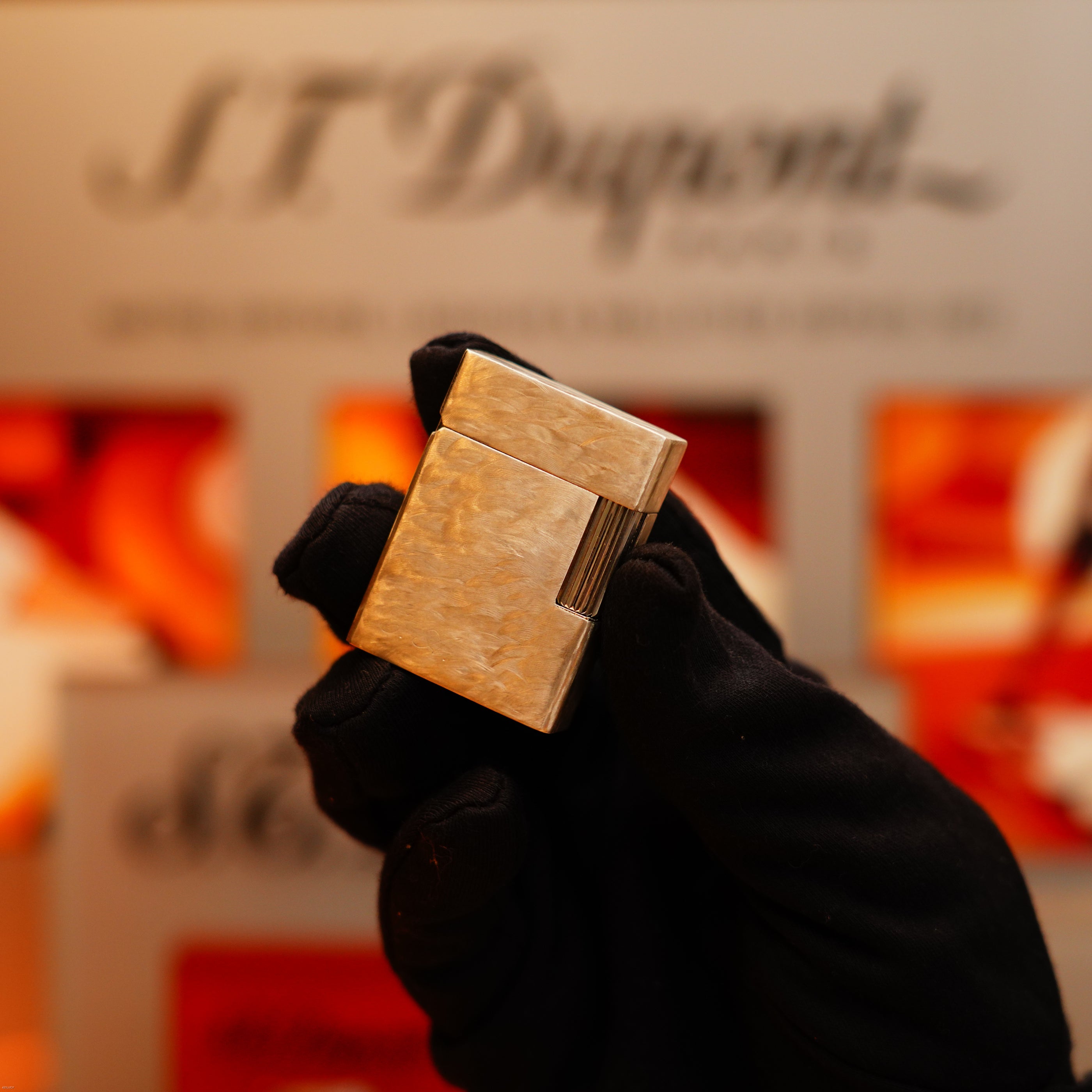 A gloved hand displays the Vintage 1970 de St Dupont Two Tone Solid 925 Sterling Silver Guilloché Pattern Lighter by S.T. Dupont against a blurred, warm-toned background with the S.T. Dupont logo visible.