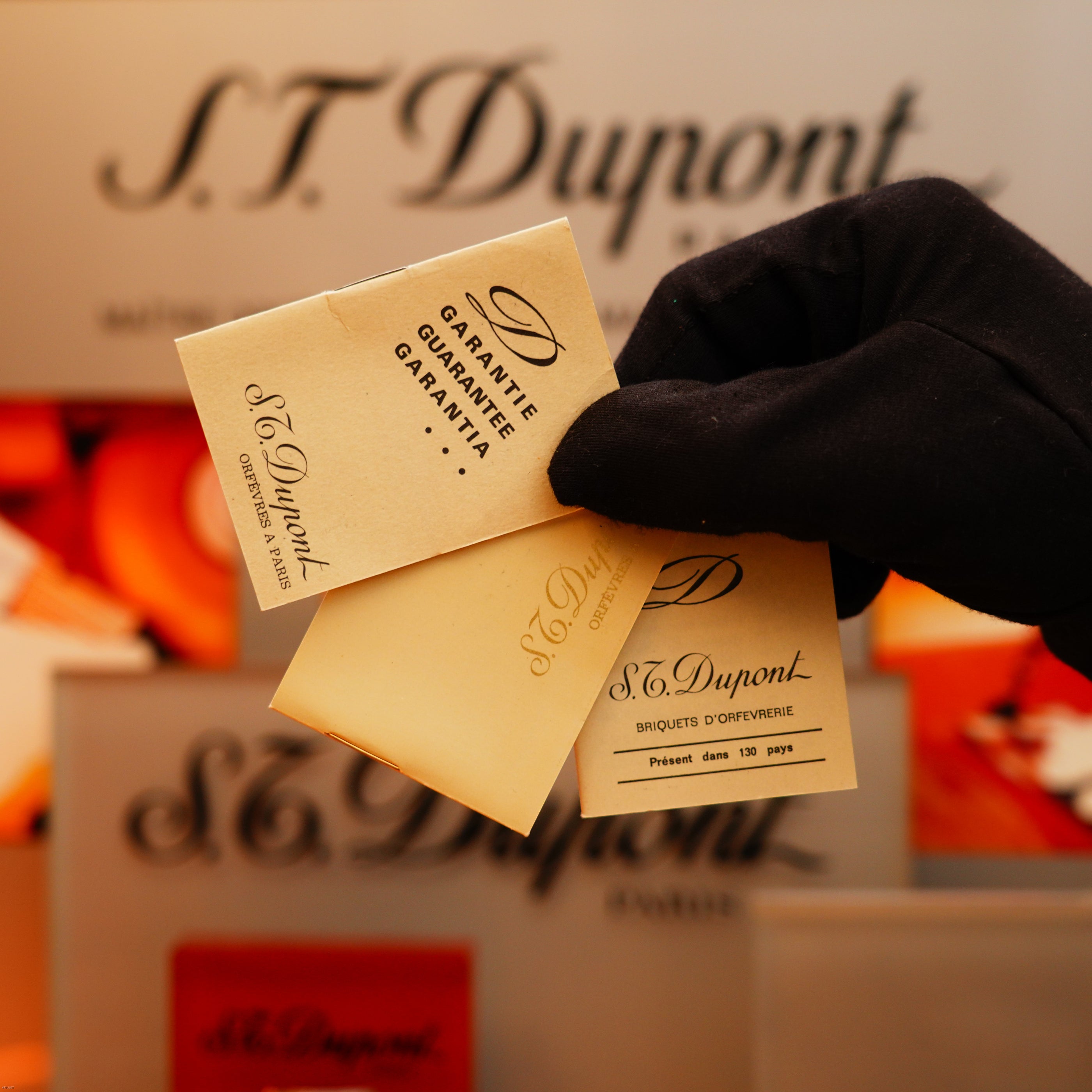 A gloved hand holds two small S.T. Dupont booklets, "Garantie" and "Briquets d'Exception," in front of a blurred background featuring the Vintage 1970 de St Dupont Two Tone Solid 925 Sterling Silver Guilloché Pattern Lighter.