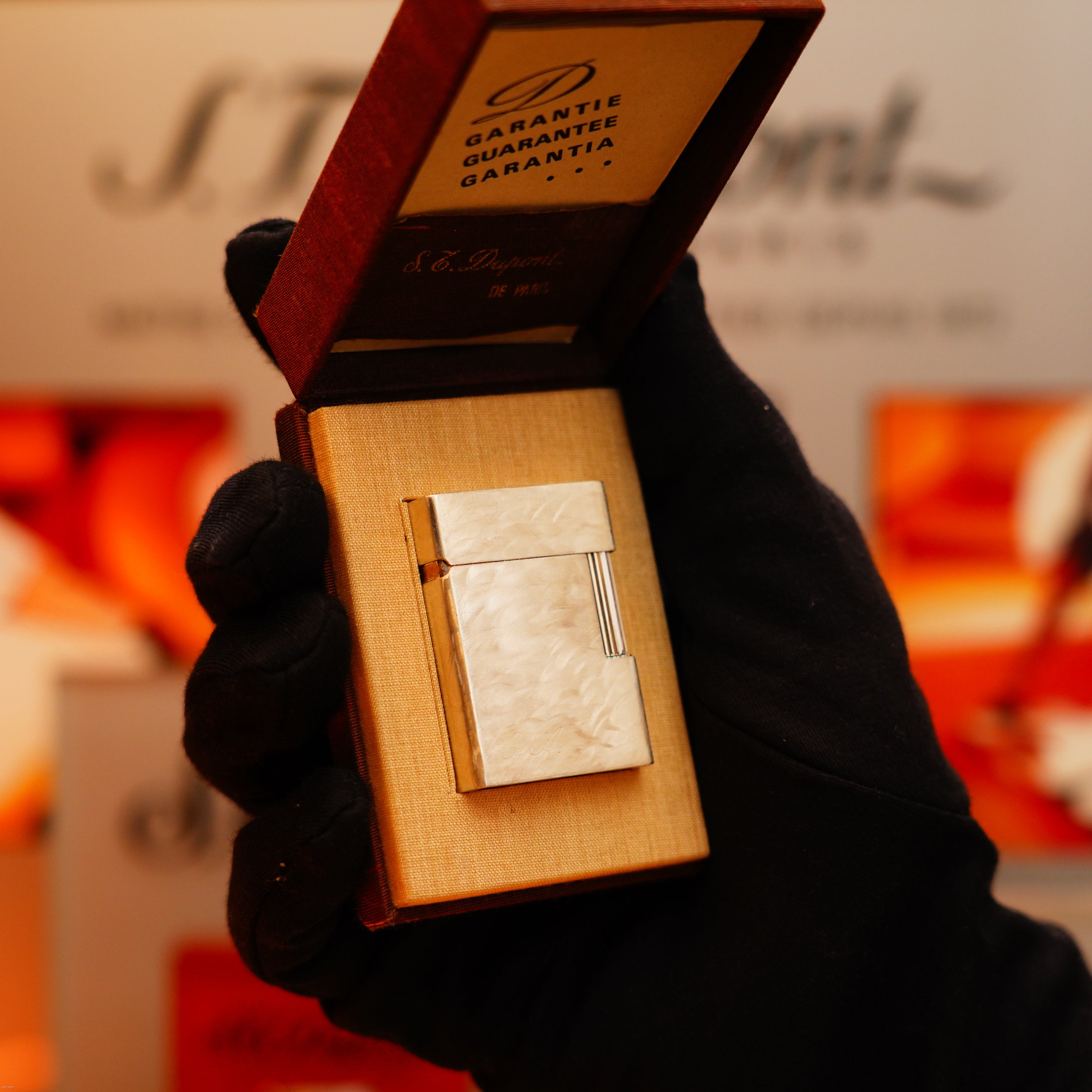 A gloved hand holds an open box with a Vintage 1970 S.T. Dupont Two Tone Solid 925 Sterling Silver Guilloché Pattern Lighter and a guarantee card inside the lid.