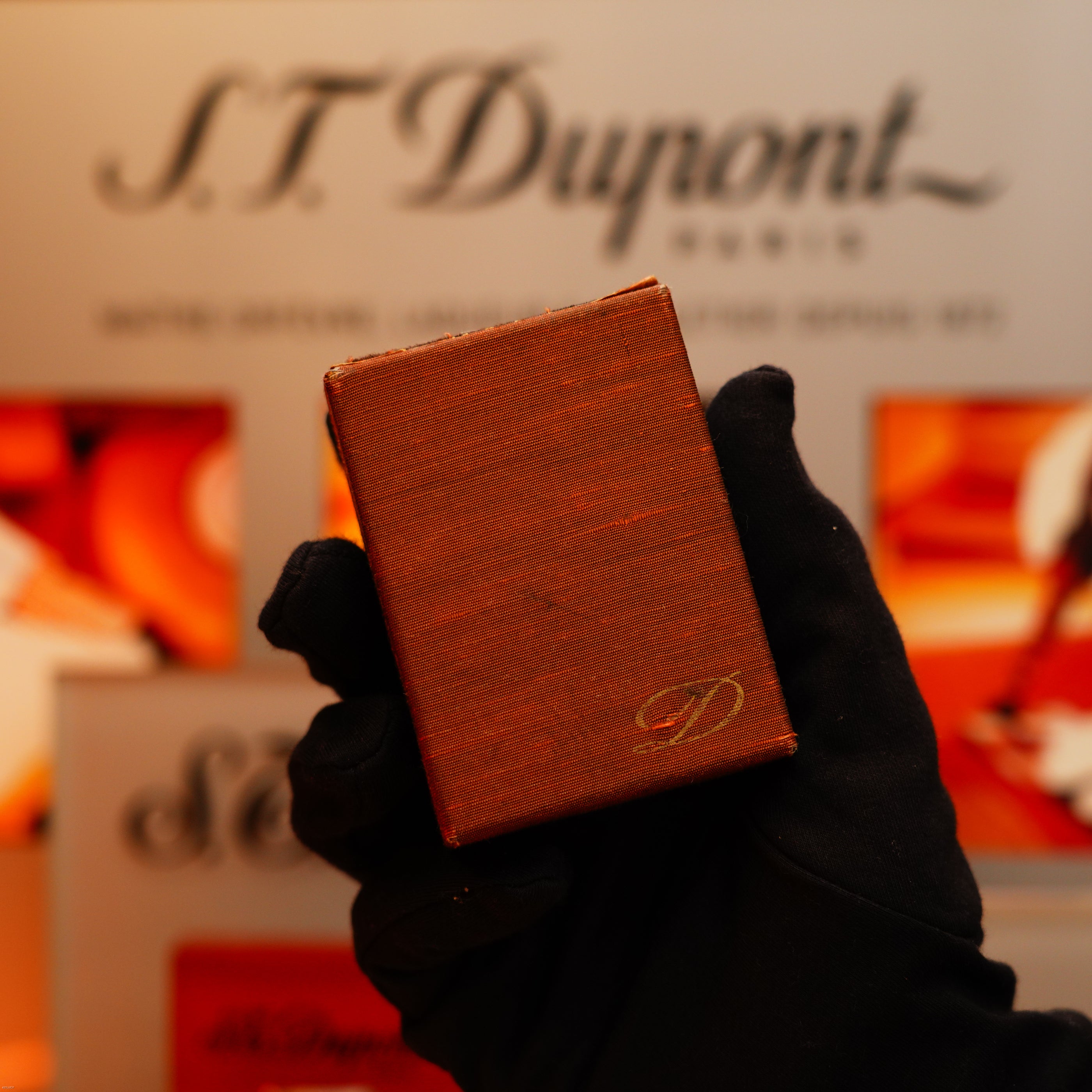 A gloved hand holds a Vintage 1970 de St Dupont Two Tone Solid 925 Sterling Silver Guilloché Pattern Lighter before an S.T. Dupont Paris display.