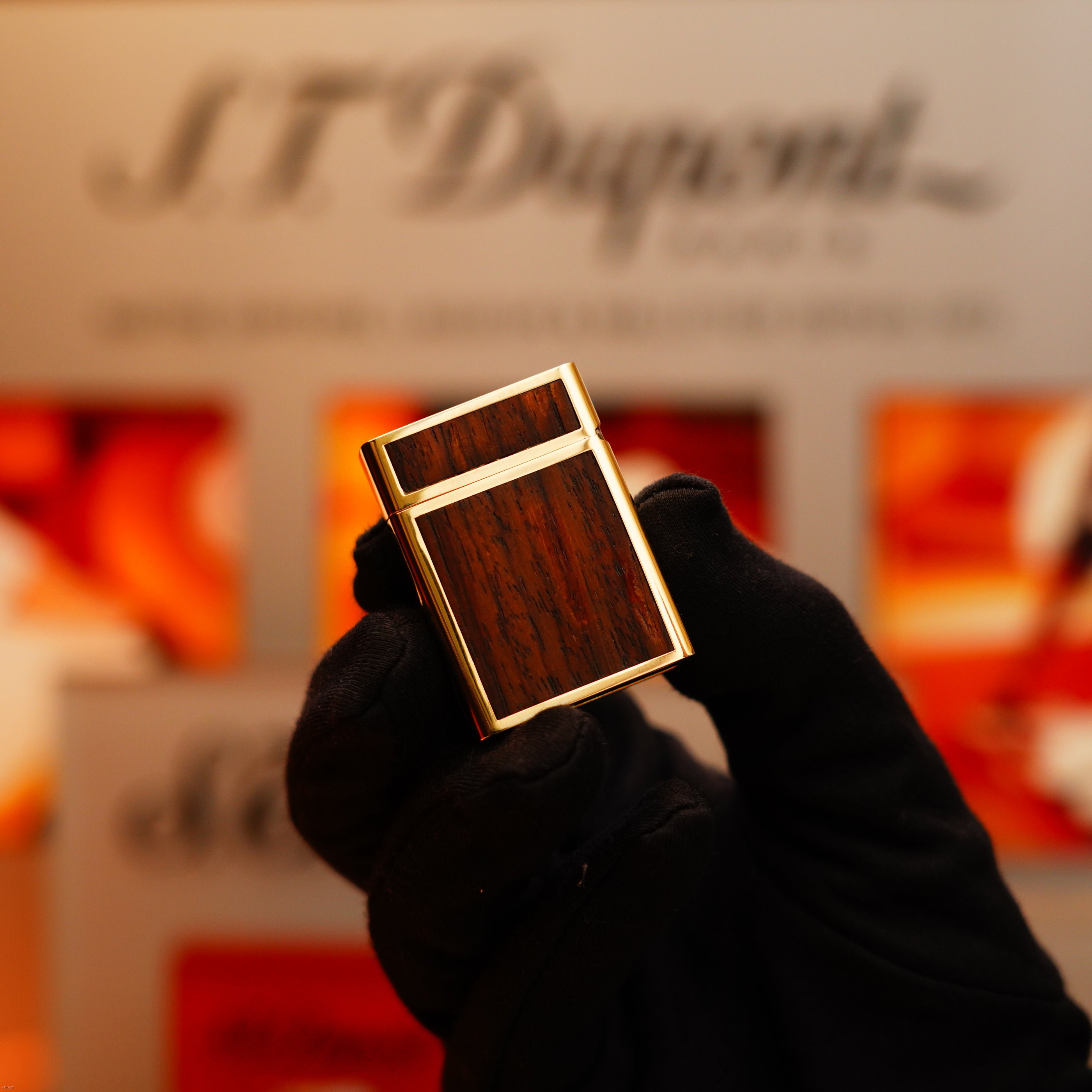 A gloved hand holds a Vintage 1970 Rare S.T. Dupont Solid 18k Gold and Natural Wood Work 750 Hallmarked Lighter against a blurred background featuring the S.T. Dupont logo.