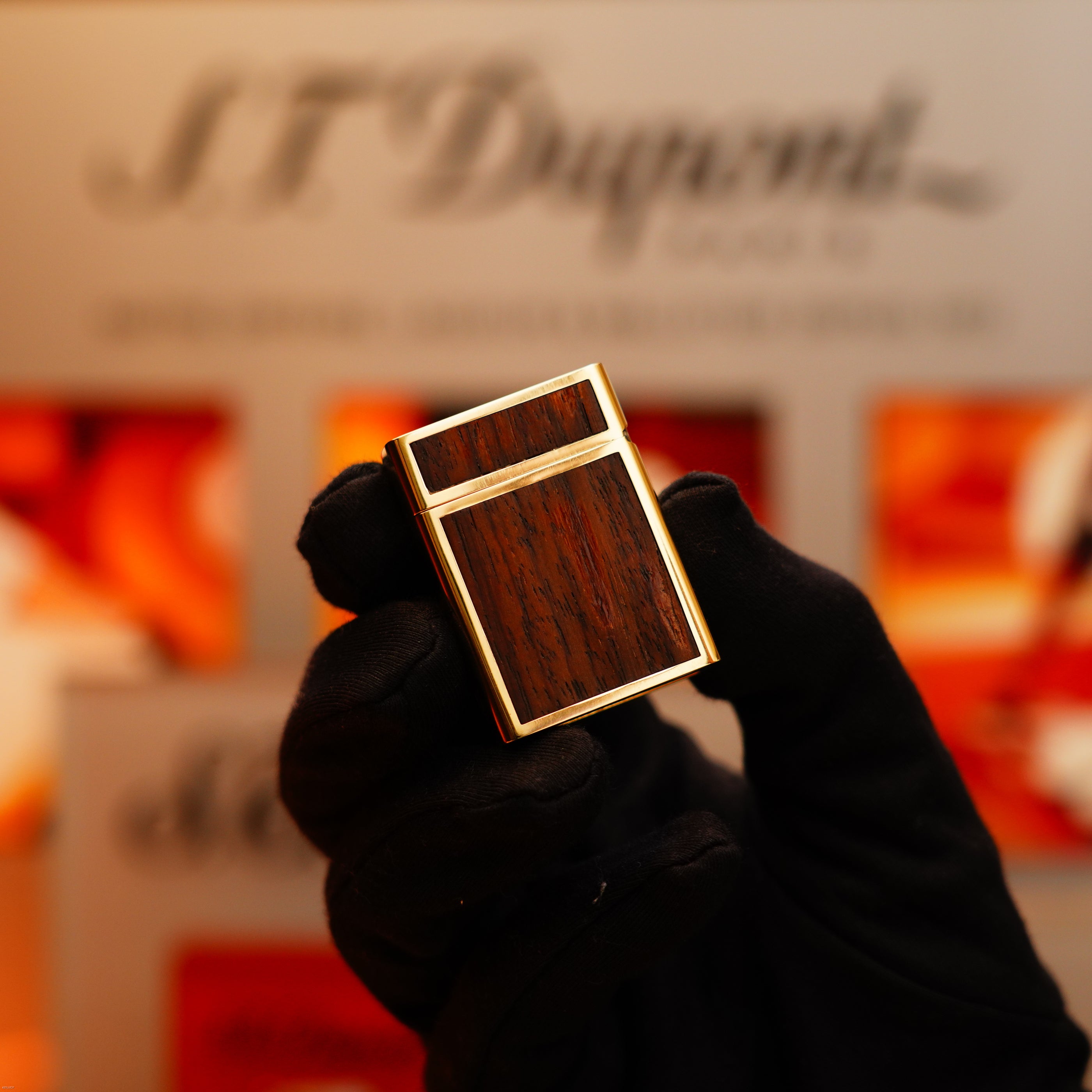 A gloved hand holds a Vintage 1970 Rare S.T. Dupont Solid 18k Gold and Natural Wood Work 750 Hallmarked Lighter before a blurred S.T. Dupont display.