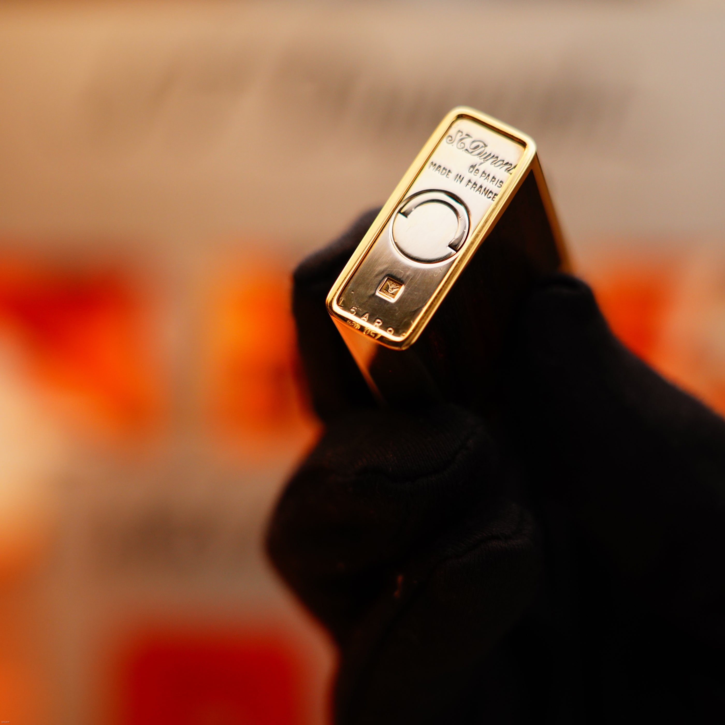 A gloved hand holds a Vintage 1970 Rare S.T. Dupont Solid 18k Gold and Natural Wood 750 hallmarked lighter, set against a blurred orange and red background.