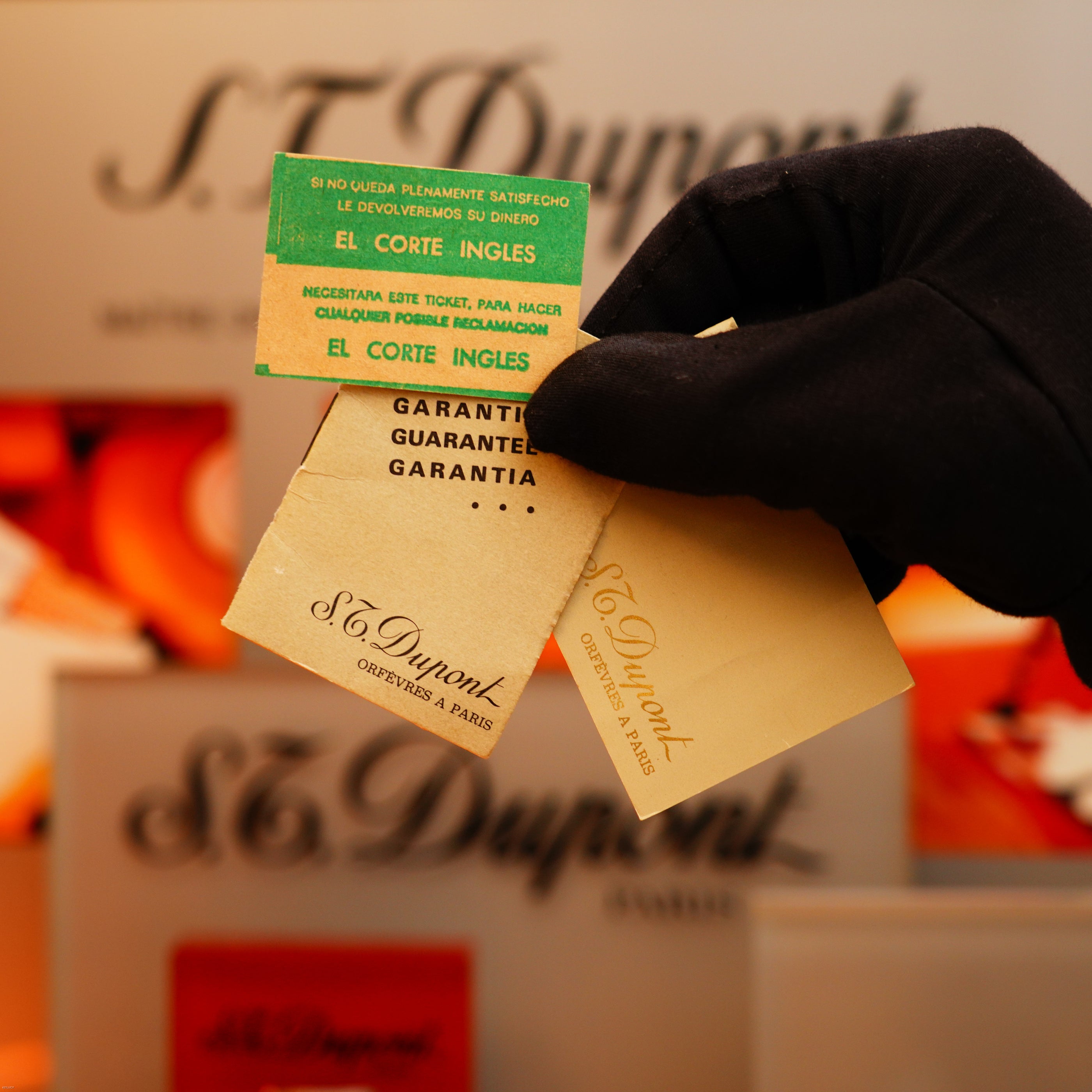 A gloved hand holds three guarantee and information cards from S.T. Dupont and El Corte Inglés, displayed against a blurred background featuring the Vintage 1970 Rare St Dupont Solid 18k Gold and Natural Wood work 750 hallmarked Lighter.
