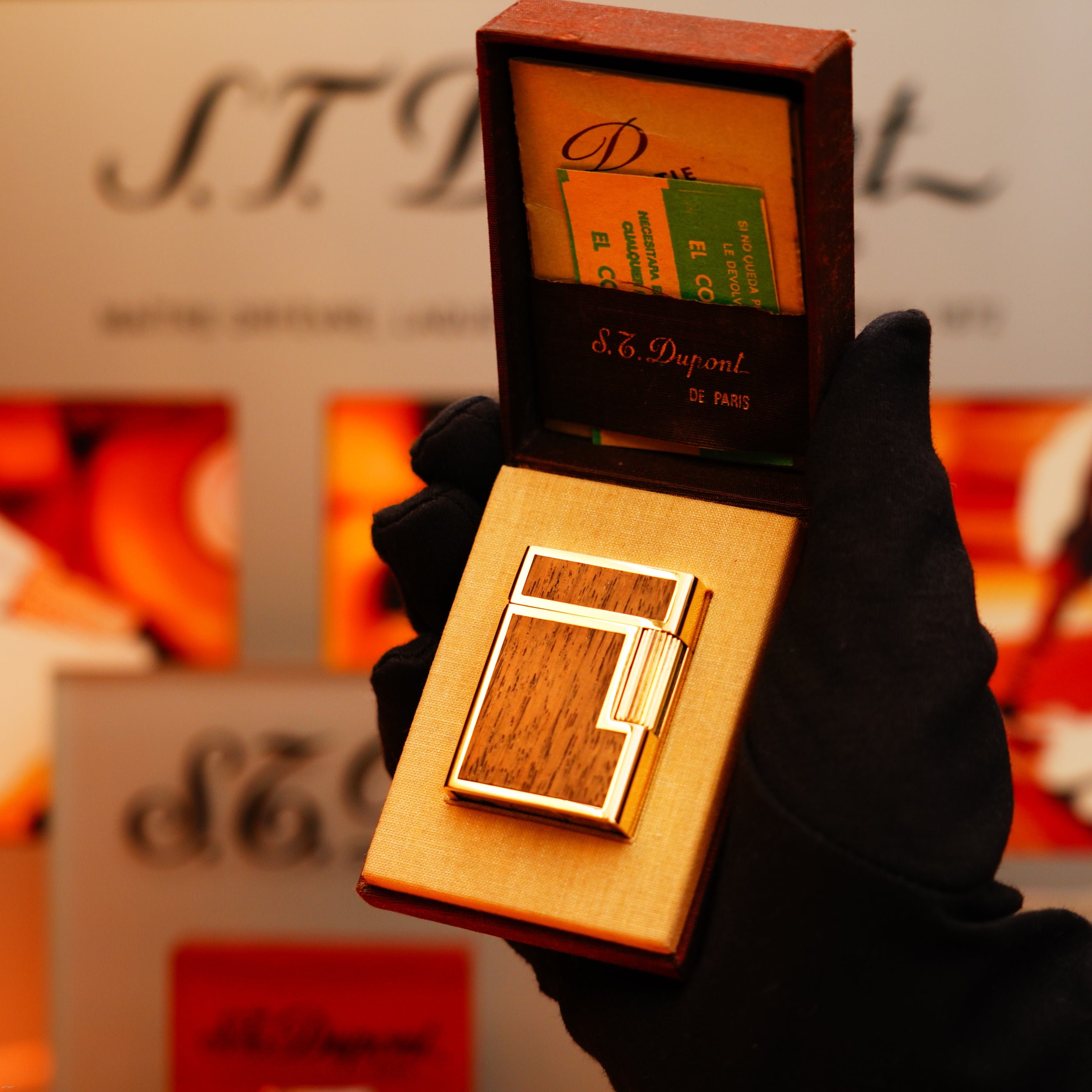 A gloved hand holds an S.T. Dupont box containing the Vintage 1970 Rare St Dupont Solid 18k Gold and Natural Woodwork 750 hallmarked Lighter, with informational cards and a blurred S.T. Dupont display in the background.