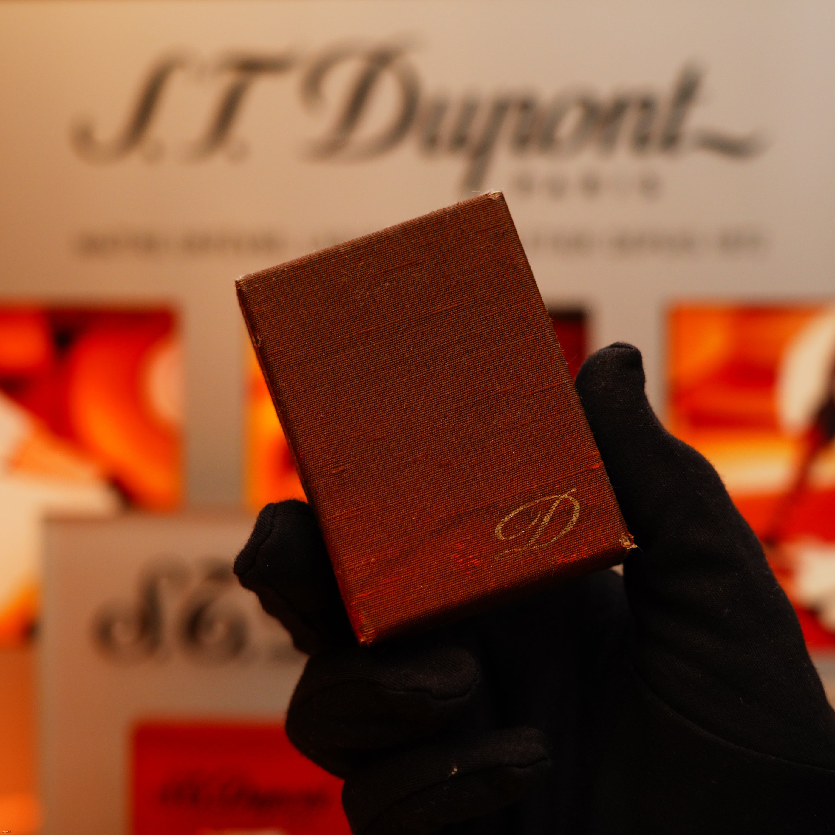 A gloved hand holds a Vintage 1970 Rare S.T. Dupont Solid 18k Gold and Natural Wood Lighter, featuring the gold "D" logo, with a blurred S.T. Dupont sign and red-orange accents in the background.