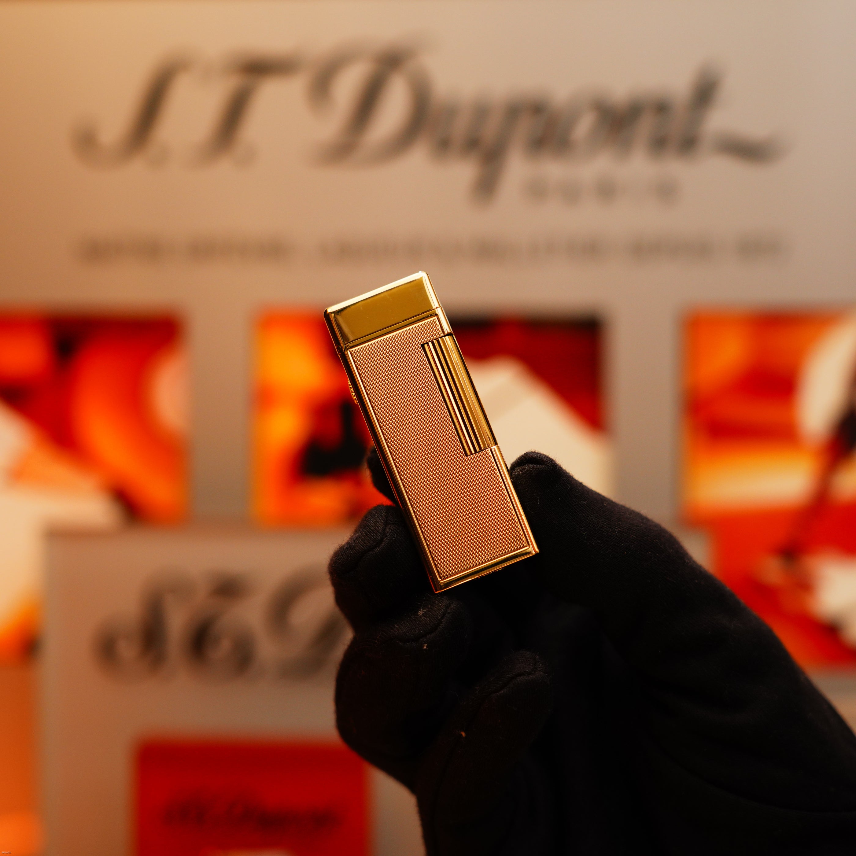 A gloved hand holds a Vintage 1964 Oct. Dunhill Rollagas Early Production Solid 18k Gold Lighter with 750 Hallmark in front of a blurred Dunhill display background.