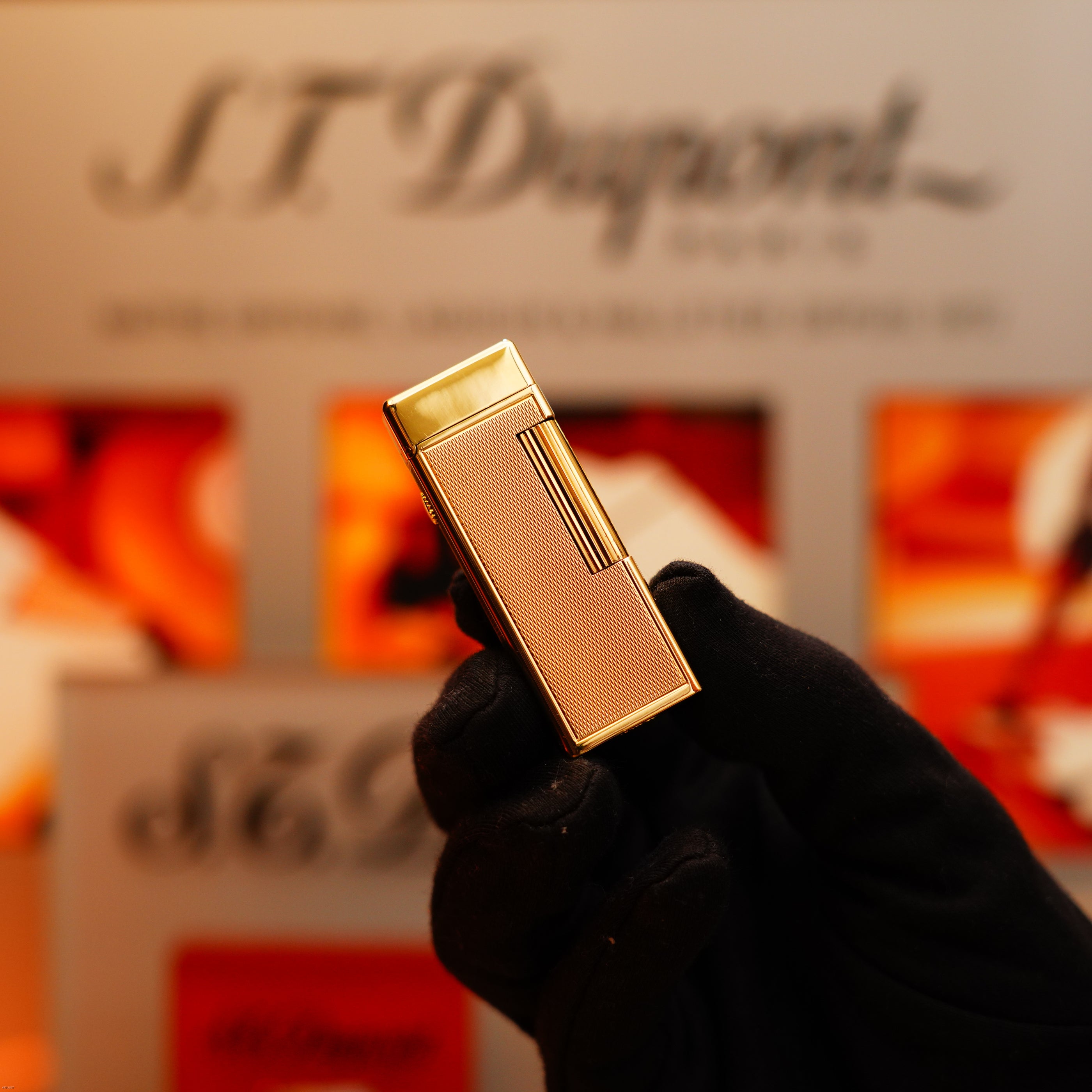 A gloved hand holds a Vintage 1964 Oct. Dunhill Rollagas Early Production Solid 18k Gold Lighter with 750 hallmark against a blurred backdrop featuring the Dunhill logo and colorful patterns.