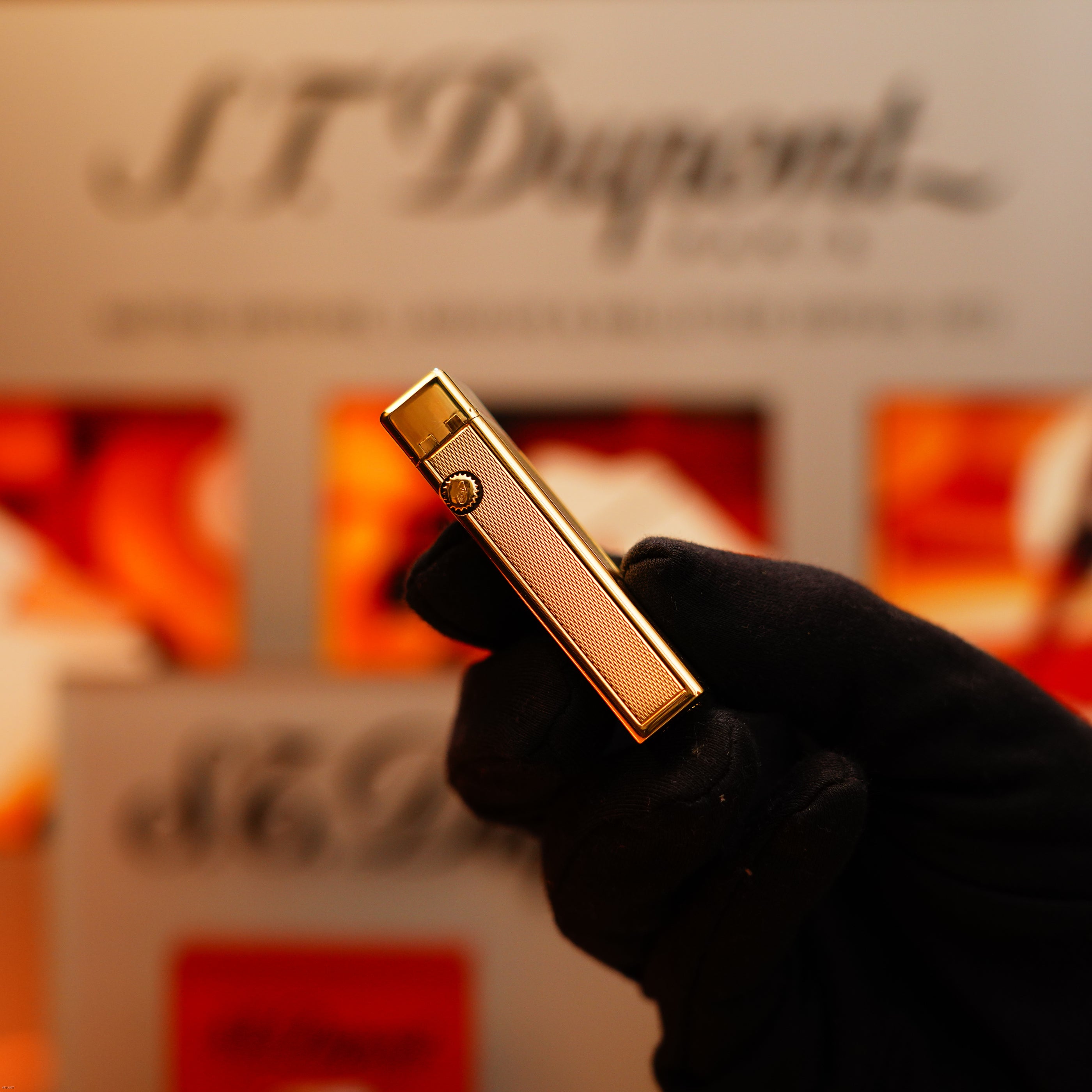 A gloved hand holds a Vintage 1964 Oct. Dunhill Rollagas Early Production Solid 18k Gold Lighter with a 750 hallmark upright, with a blurred Dunhill display in the background.
