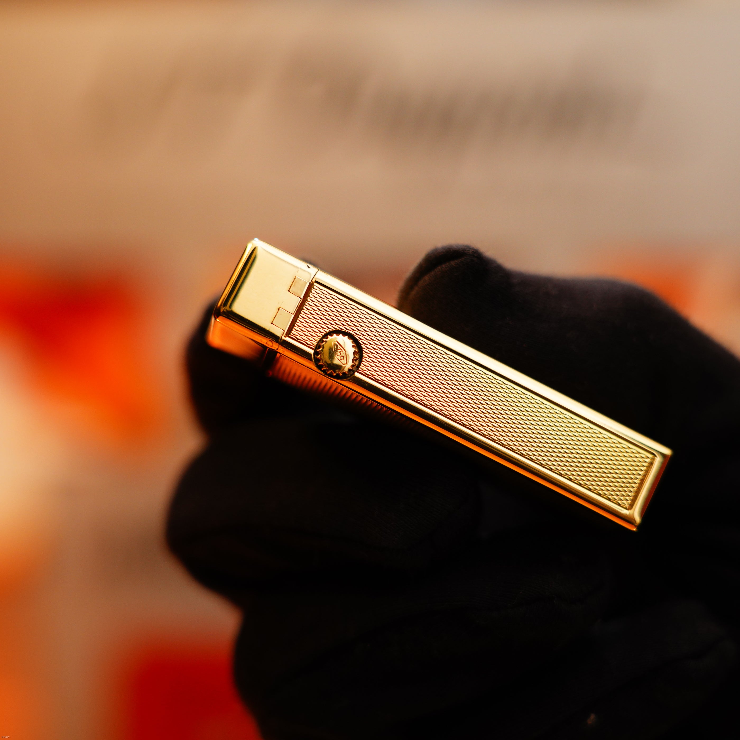 A gloved hand holds a Dunhill Vintage 1964 Oct. Rollagas Early Production Solid 18k Gold Lighter (750 Hallmark, Extremely Heavy) against a blurred orange and white background.