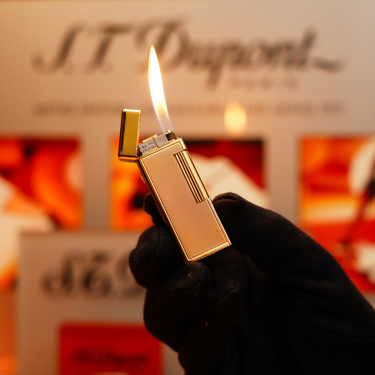 A gloved hand holds a vintage 1964 Dunhill Rollagas lighter in solid 18k gold, its flame lit, set against a blurred branded background.