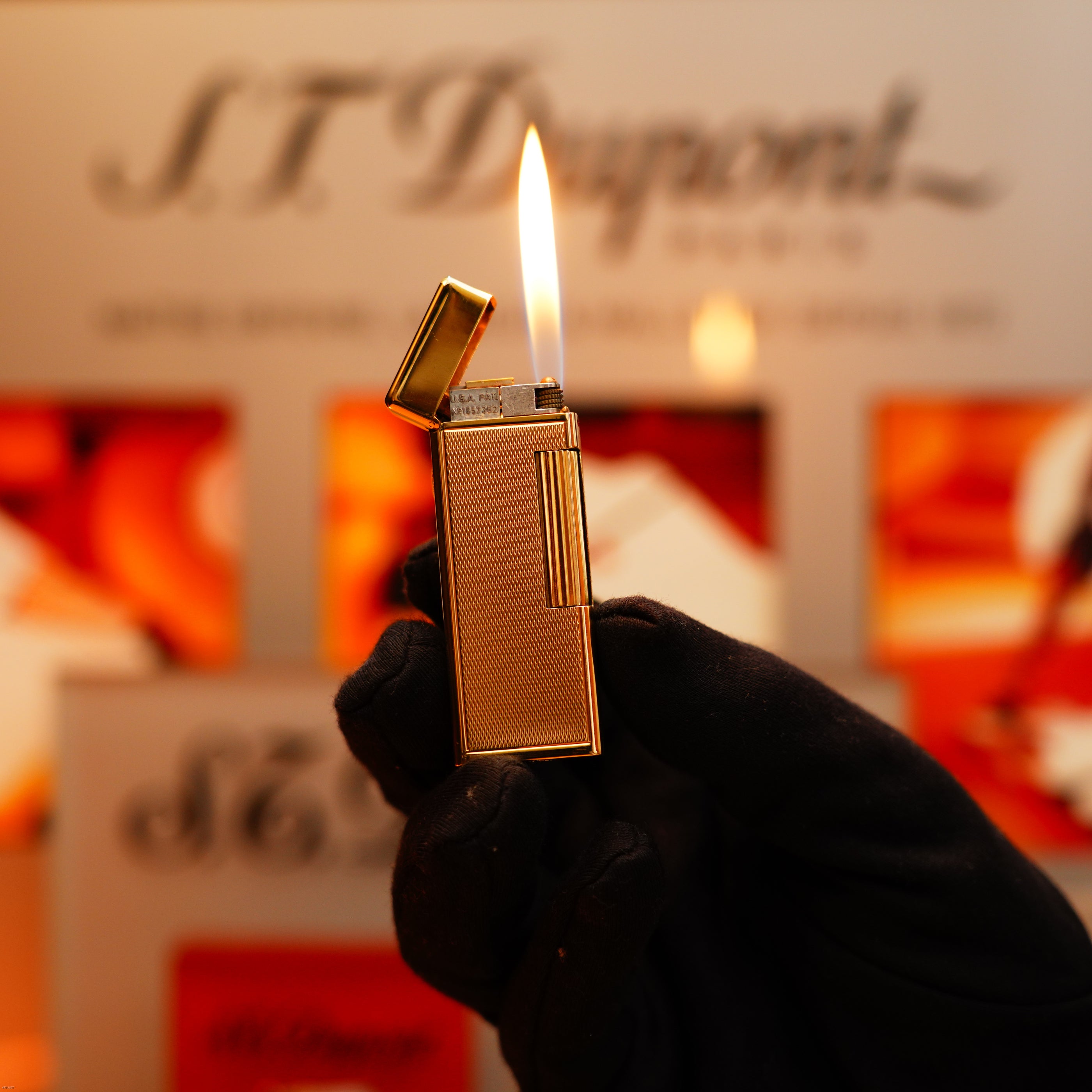 A gloved hand holds a Vintage 1964 Oct. Dunhill Rollagas Early Production Solid 18k Gold Lighter with a lit flame, its 750 hallmark visible, against a blurred background featuring the Dunhill logo.