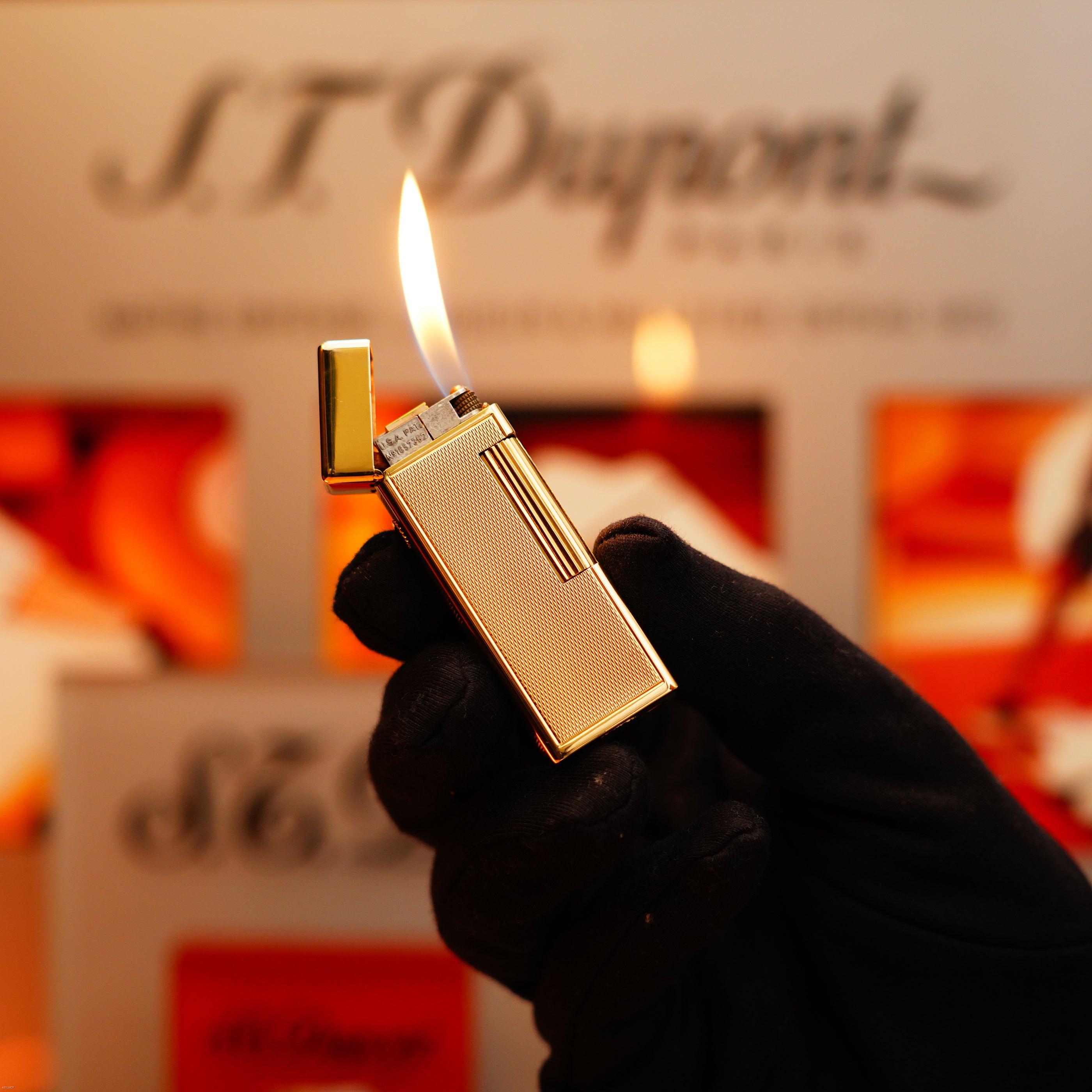 A gloved hand holds a lit Vintage 1964 Oct. Dunhill Rollagas Early Production Solid 18k Gold Lighter with 750 Hallmark against a blurred background featuring the Dunhill logo.