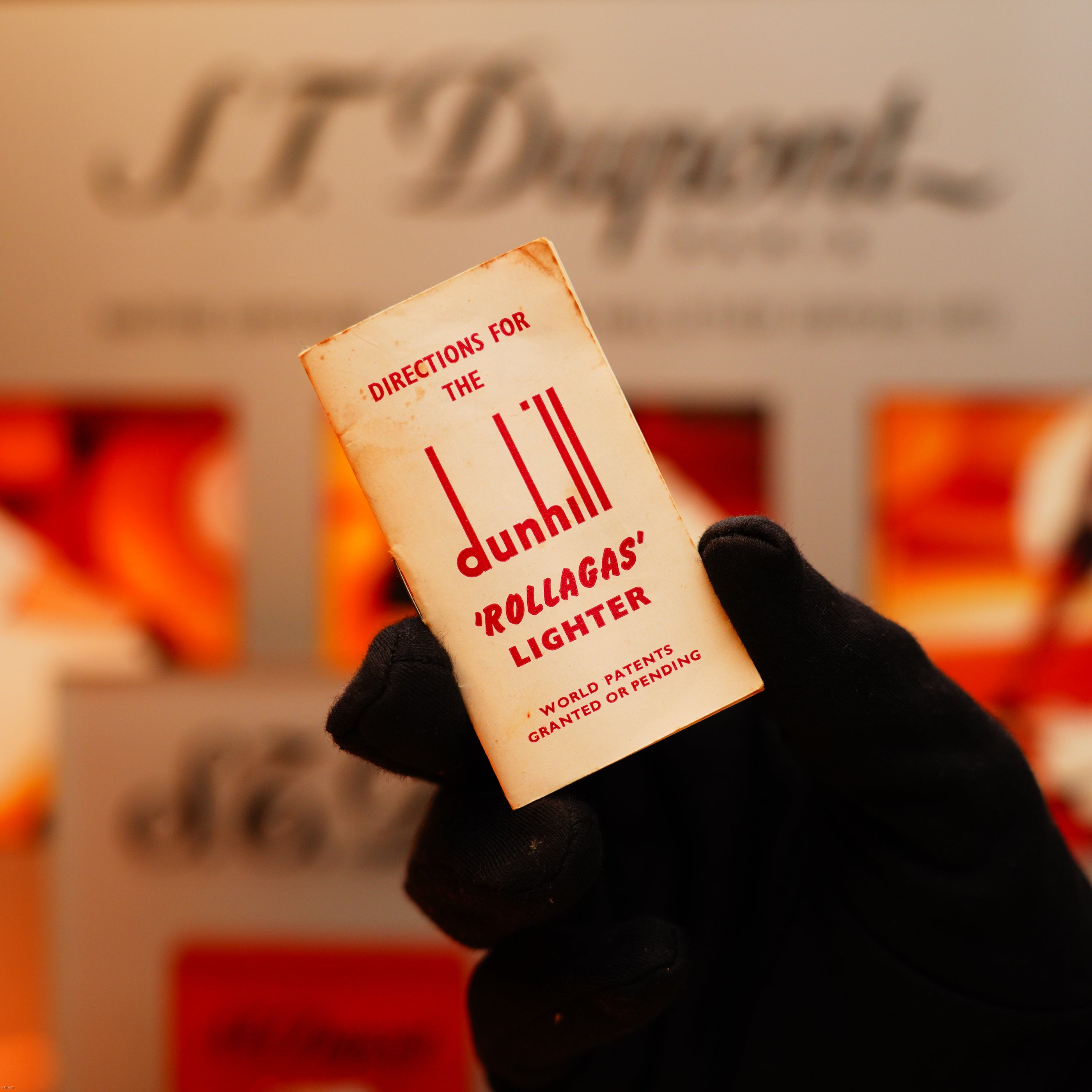 A gloved hand holds a small instruction booklet for the Dunhill Vintage 1964 Oct. Rollagas Early Production Solid 18k Gold Lighter, with blurred Dunhill-branded signage in the background.