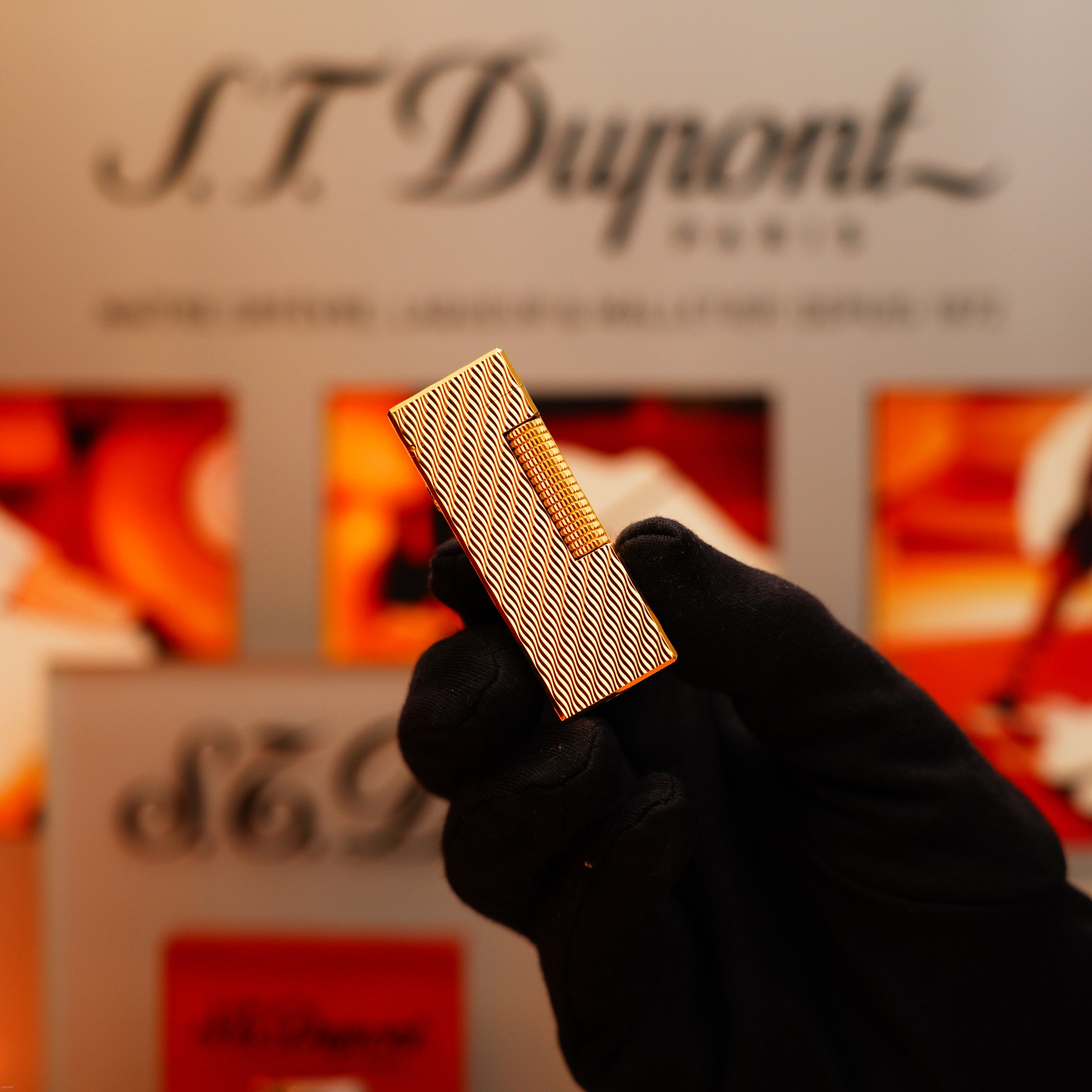 A gloved hand holds a Vintage 1971 Dunhill 24k Gold Finish Rollagas Lighter with a rare guilloché wave pattern, set against a blurred background featuring the Dunhill logo.