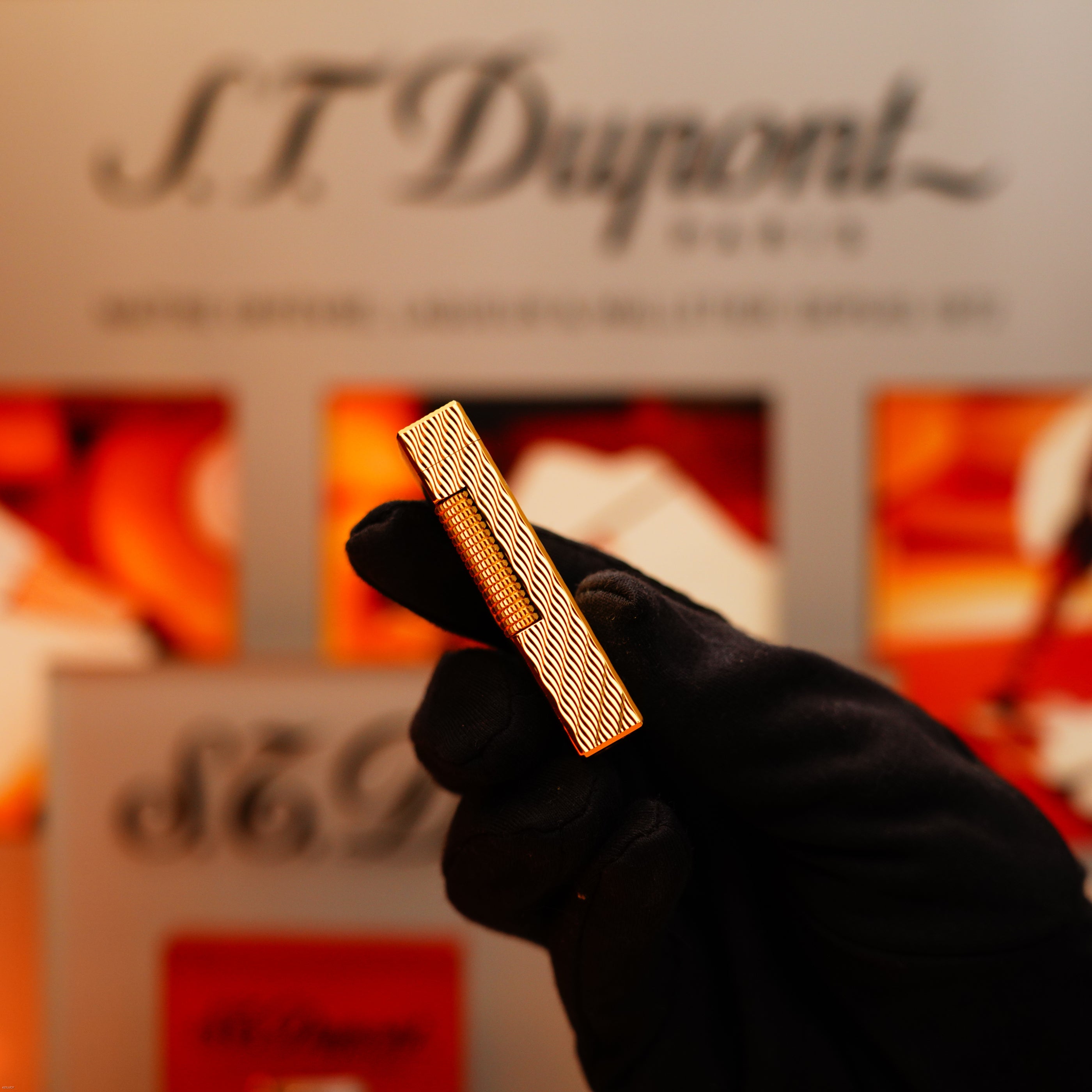 A gloved hand holds a vintage 1971 Dunhill Rollagas lighter with a rare 24k gold finish and guilloché wave pattern, with a blurred Dunhill display in the background.