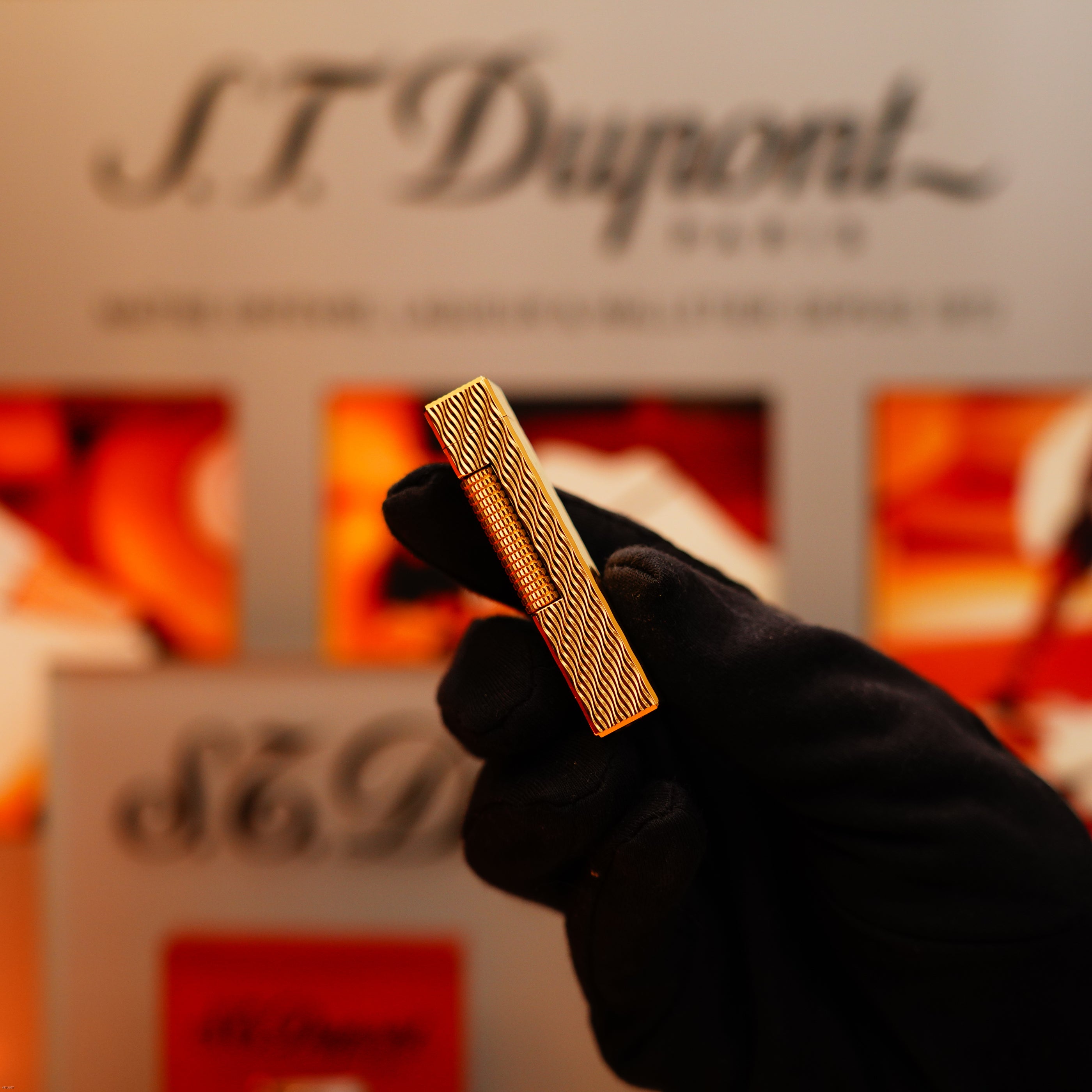 A gloved hand holds a Vintage 1971 Dunhill 24k Gold Finish Rollagas Lighter with Rare Guilloché Wave Pattern in front of a blurred S.T. Dupont display.