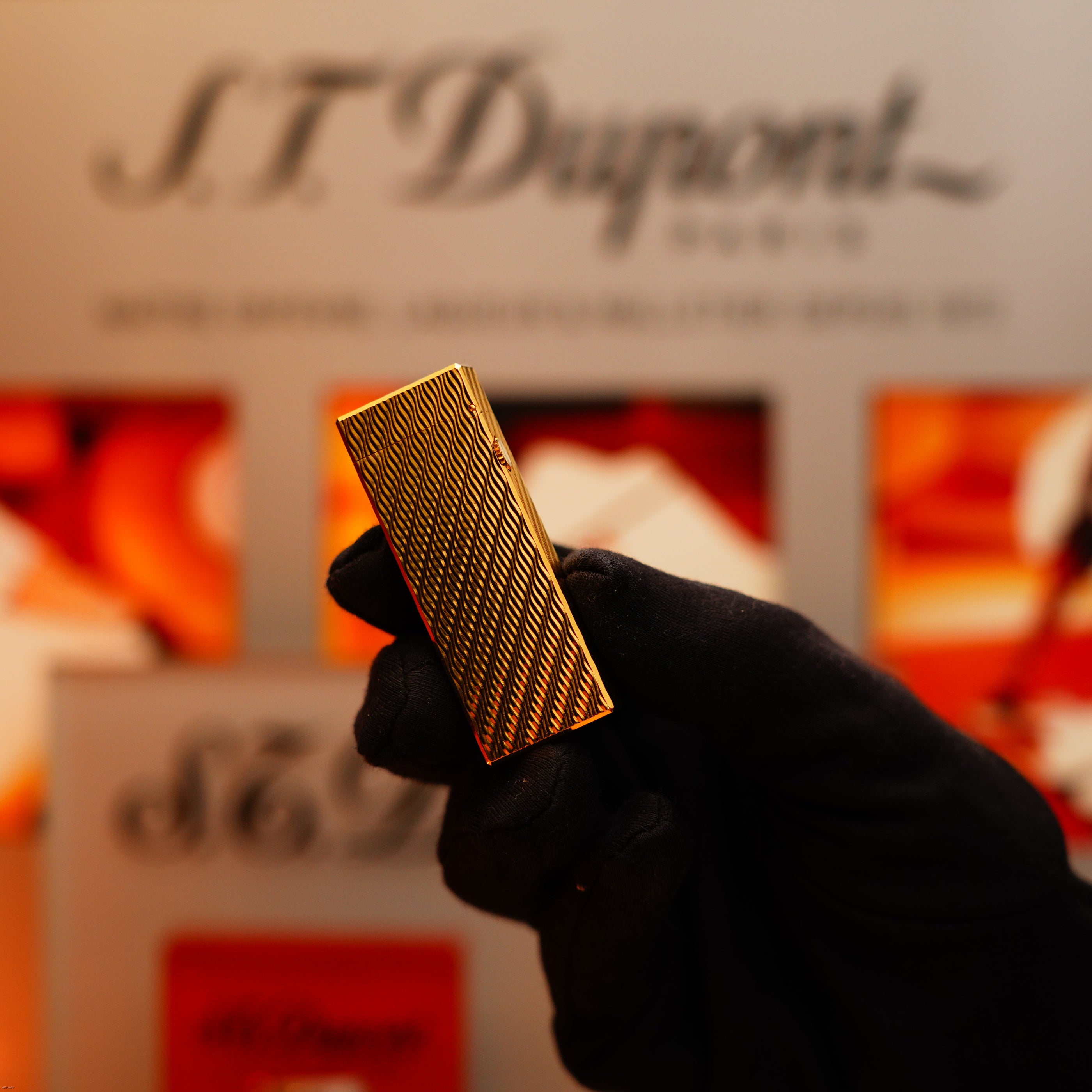 A gloved hand holds a Vintage 1971 Dunhill 24k Gold Finish Rollagas lighter with a rare guilloché wave pattern, set against a blurred branded background.
