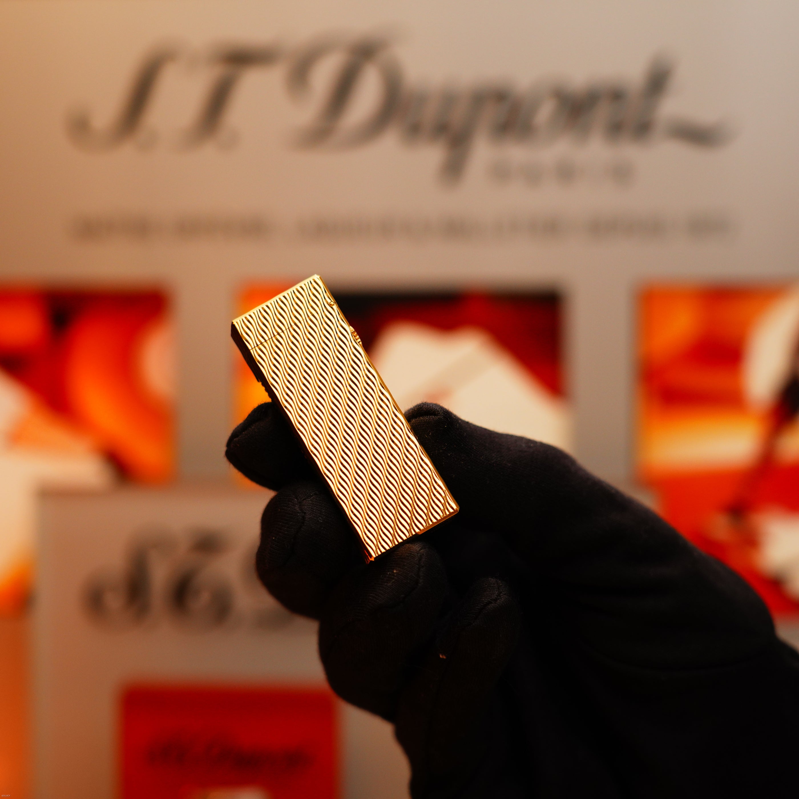 A gloved hand holds a Vintage 1971 Dunhill 24k Gold Finish Rollagas Guilloché Wave Pattern lighter, with a blurred Dunhill sign in the background.