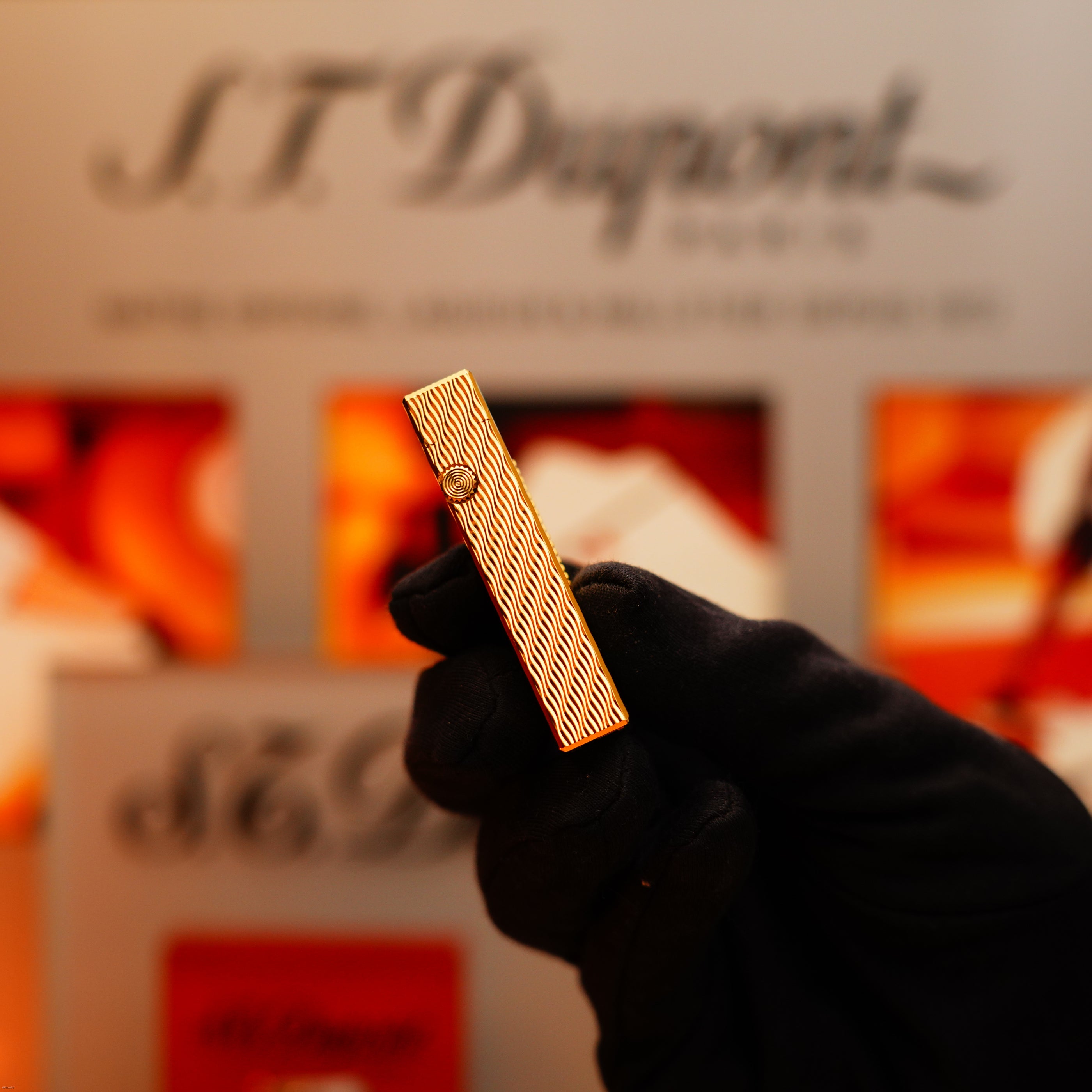 A gloved hand holds a Vintage 1971 Dunhill 24k Gold Finish Rollagas Rare Guilloché Wave Pattern Initial Version Lighter in front of a blurred branded display background.