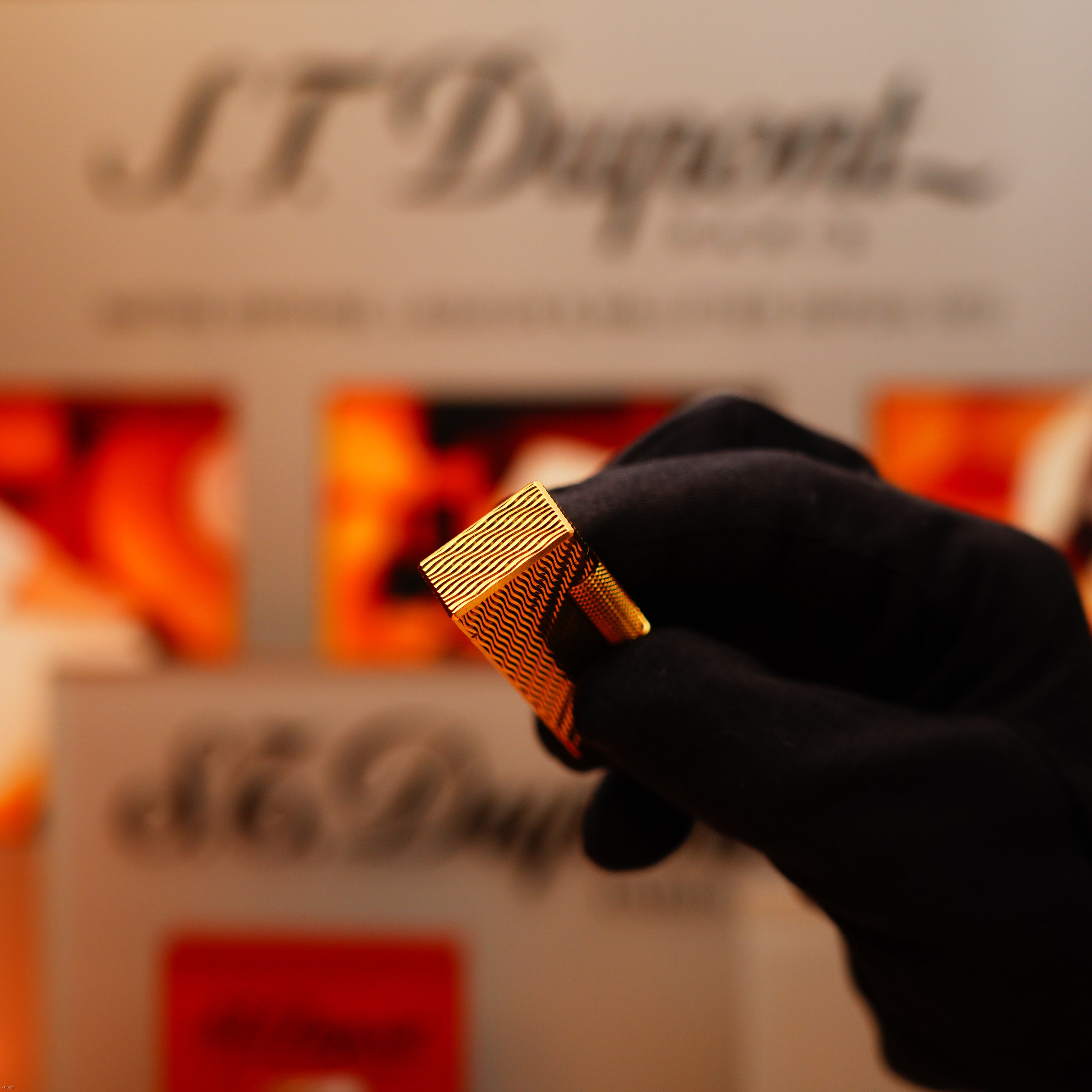 A gloved hand holds a Vintage 1971 Dunhill 24k Gold Finish Rollagas Lighter with rare guilloché wave pattern; blurred Dunhill packaging appears in the background.