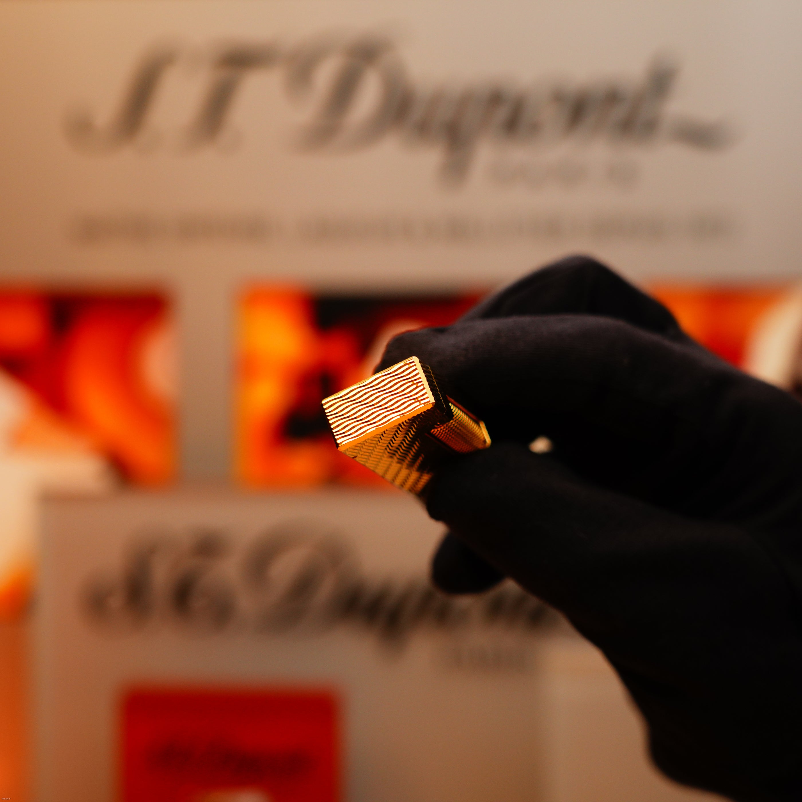 A gloved hand holds a Vintage 1971 Dunhill 24k Gold Finish Rollagas Guilloché Wave Pattern lighter, in front of a blurred display.
