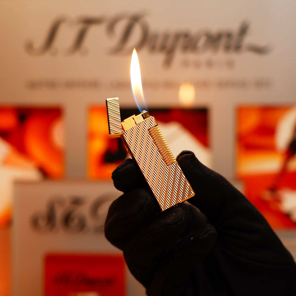 A gloved hand holds a lit Vintage 1971 Dunhill 24k Gold Finish Rollagas Rare Guilloché Wave Pattern Lighter before a blurred display background.