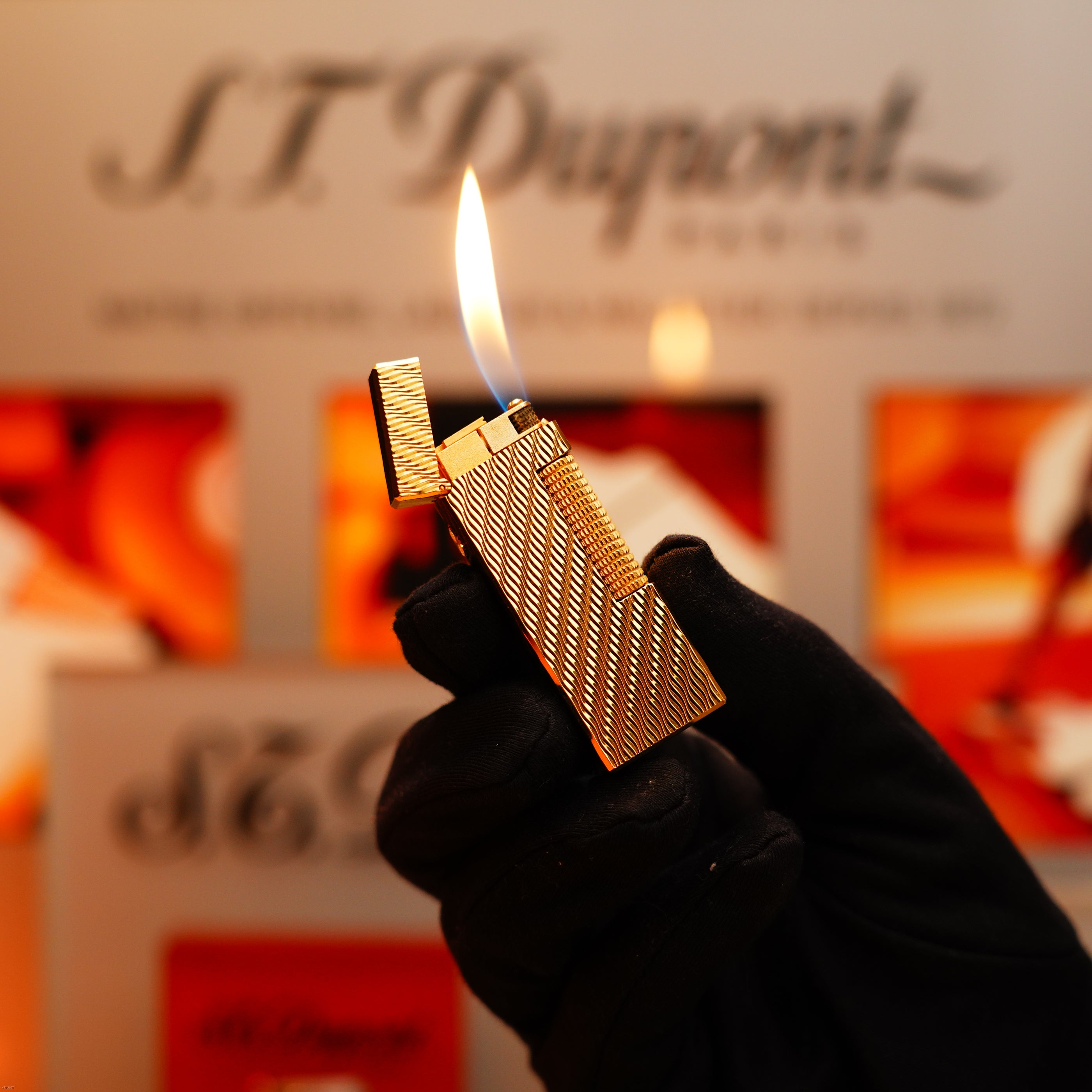 A gloved hand holds an open Vintage 1971 Dunhill 24k Gold Finish Rollagas Lighter with flame, featuring a rare Guilloché Wave pattern, in front of an out-of-focus S.T. Dupont sign and red-orange background.
