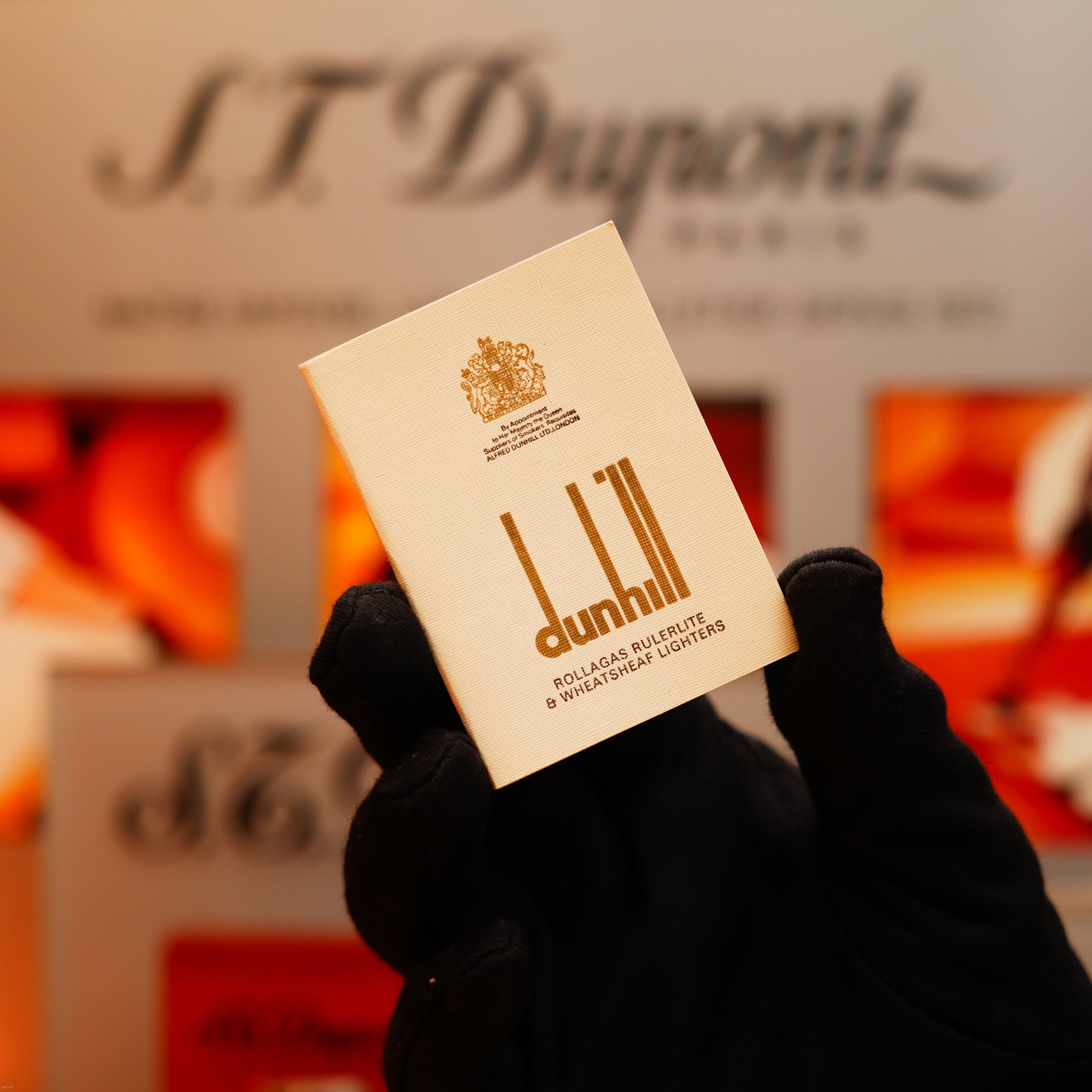 A gloved hand holds a Vintage 1971 Dunhill 24k Gold Finish Rollagas Rare Visual Guilloché Wave Pattern Initial Version Lighter in front of a blurred ST Dupont display.