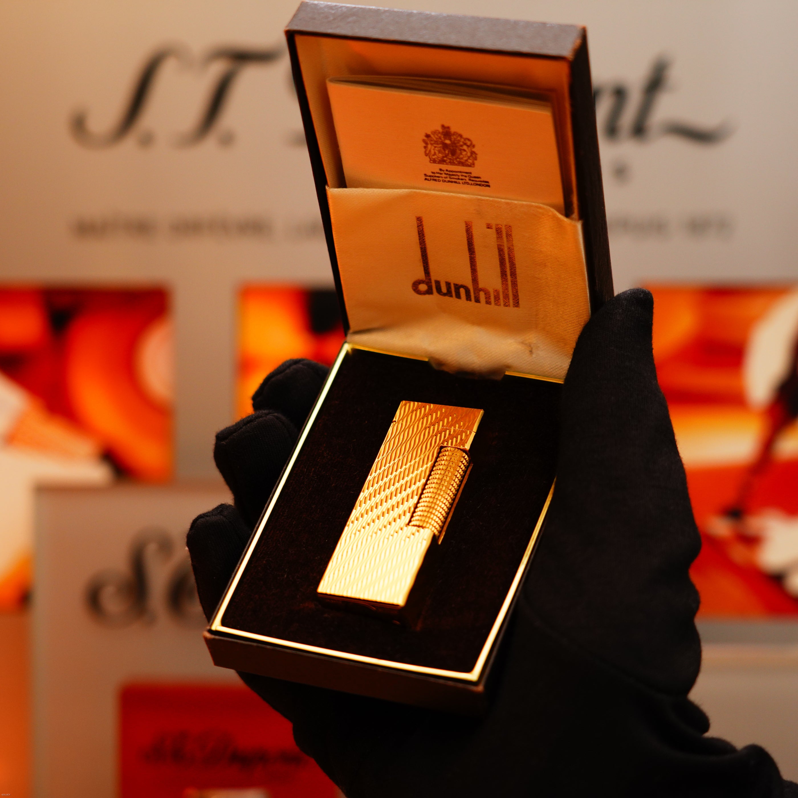 A gloved hand holds an open Dunhill box displaying a Vintage 1971 Dunhill 24k Gold Finish Rollagas Rare Visual Guilloché Wave Pattern Initial Version Lighter, set against a blurred background.
