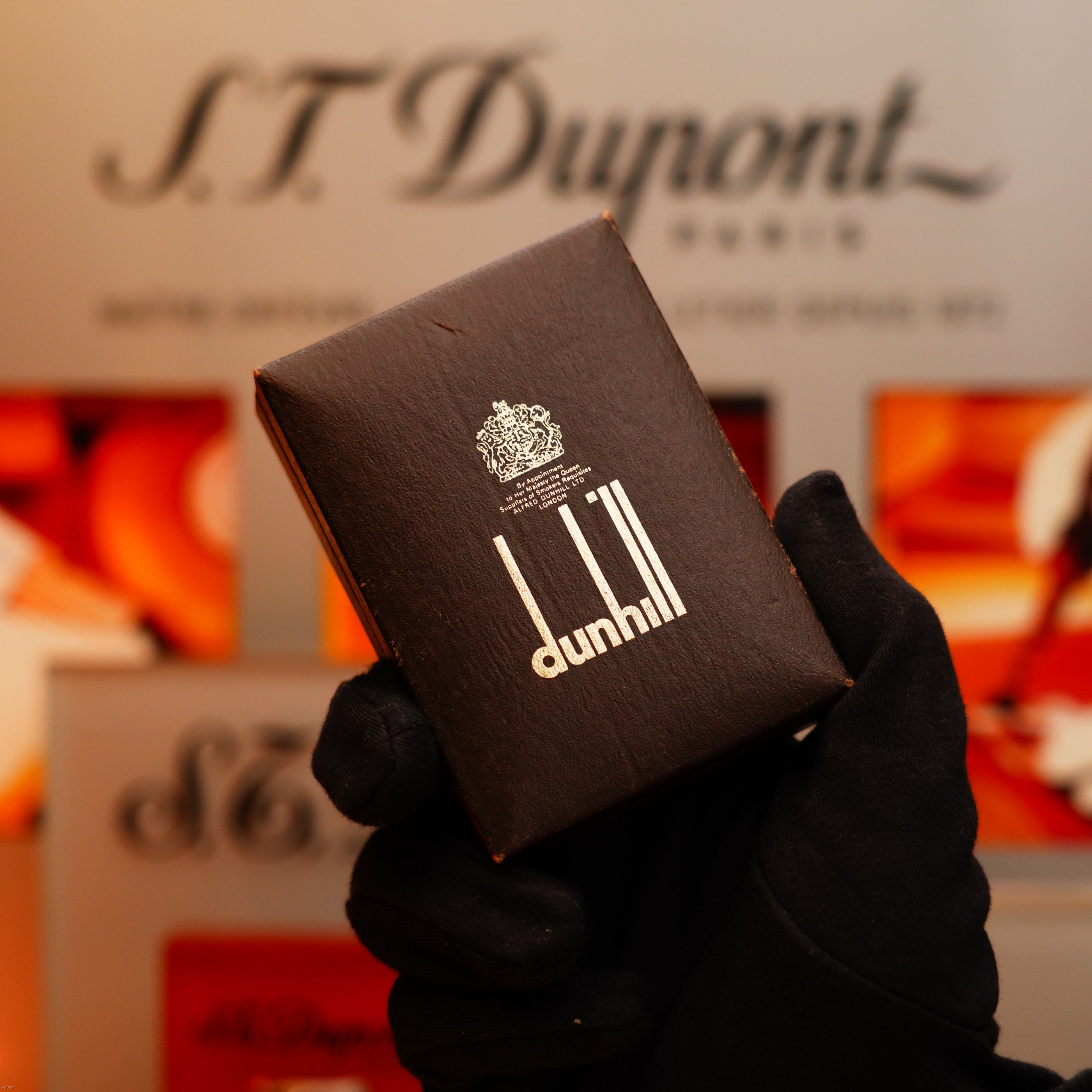 A gloved hand holds a Vintage 1971 Dunhill 24k Gold Finish Rollagas Lighter with Guilloché wave pattern, in front of a blurred S.T. Dupont Paris display.