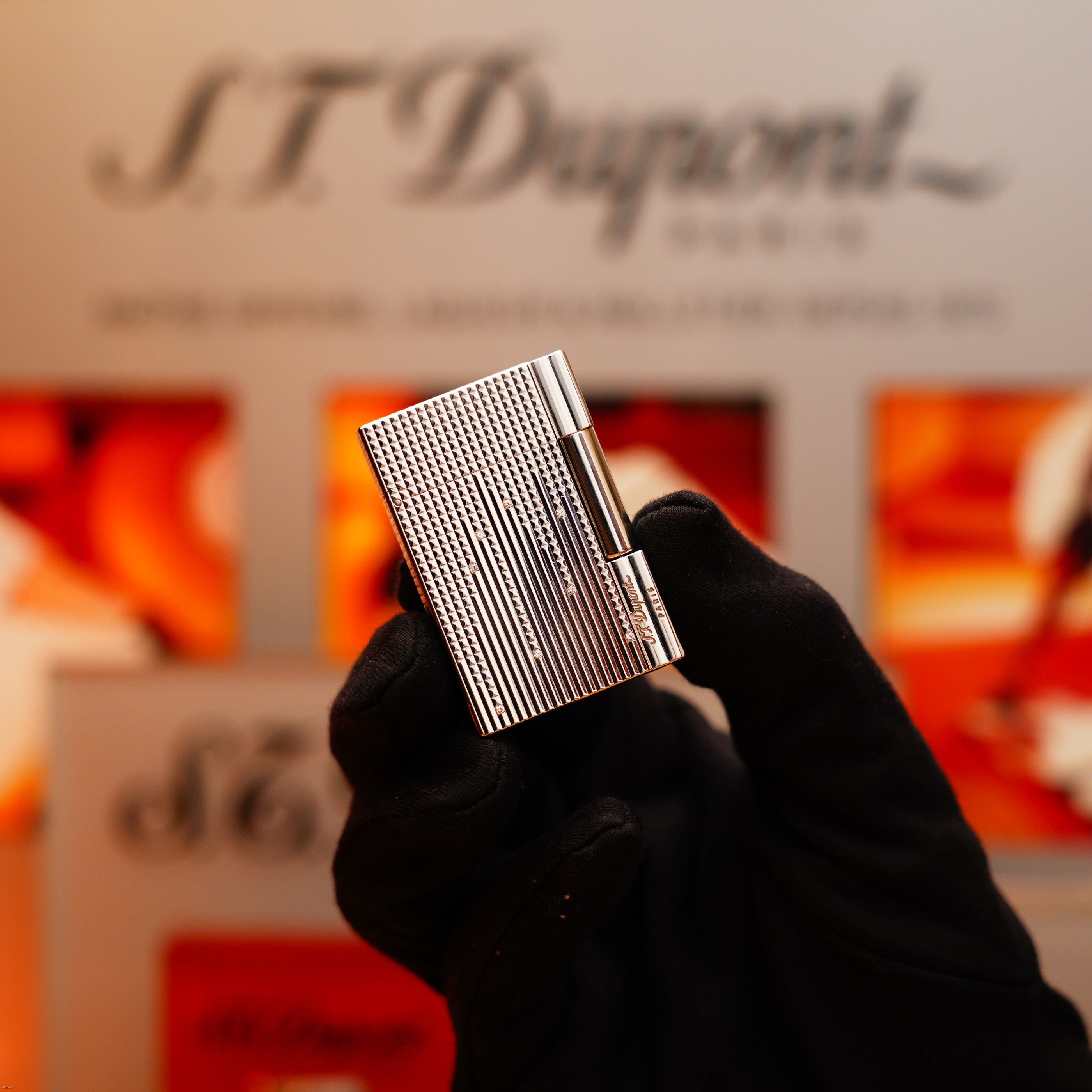 A gloved hand holds a Vintage 2005 S.T. Dupont Gatsby Diamond Drops Limited Edition Lighter with 16 natural diamonds and heavy platinum, in front of a blurred background featuring the S.T. Dupont logo and branding.