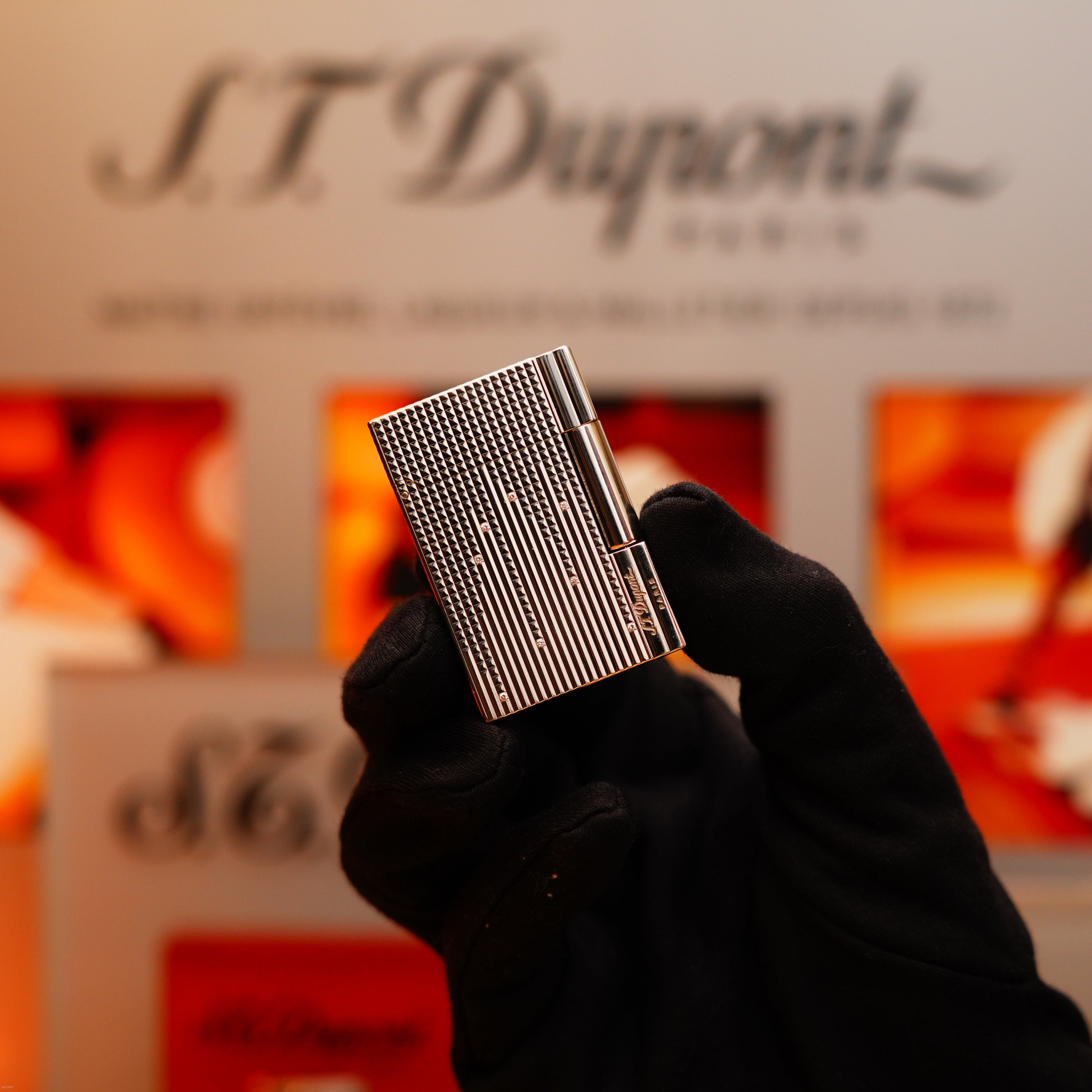 A gloved hand holds the Vintage 2005 S.T. Dupont Gatsby Diamond Drops Limited Edition Lighter with 16 natural diamonds and heavy platinum, displayed before a blurred background featuring the S.T. Dupont logo.