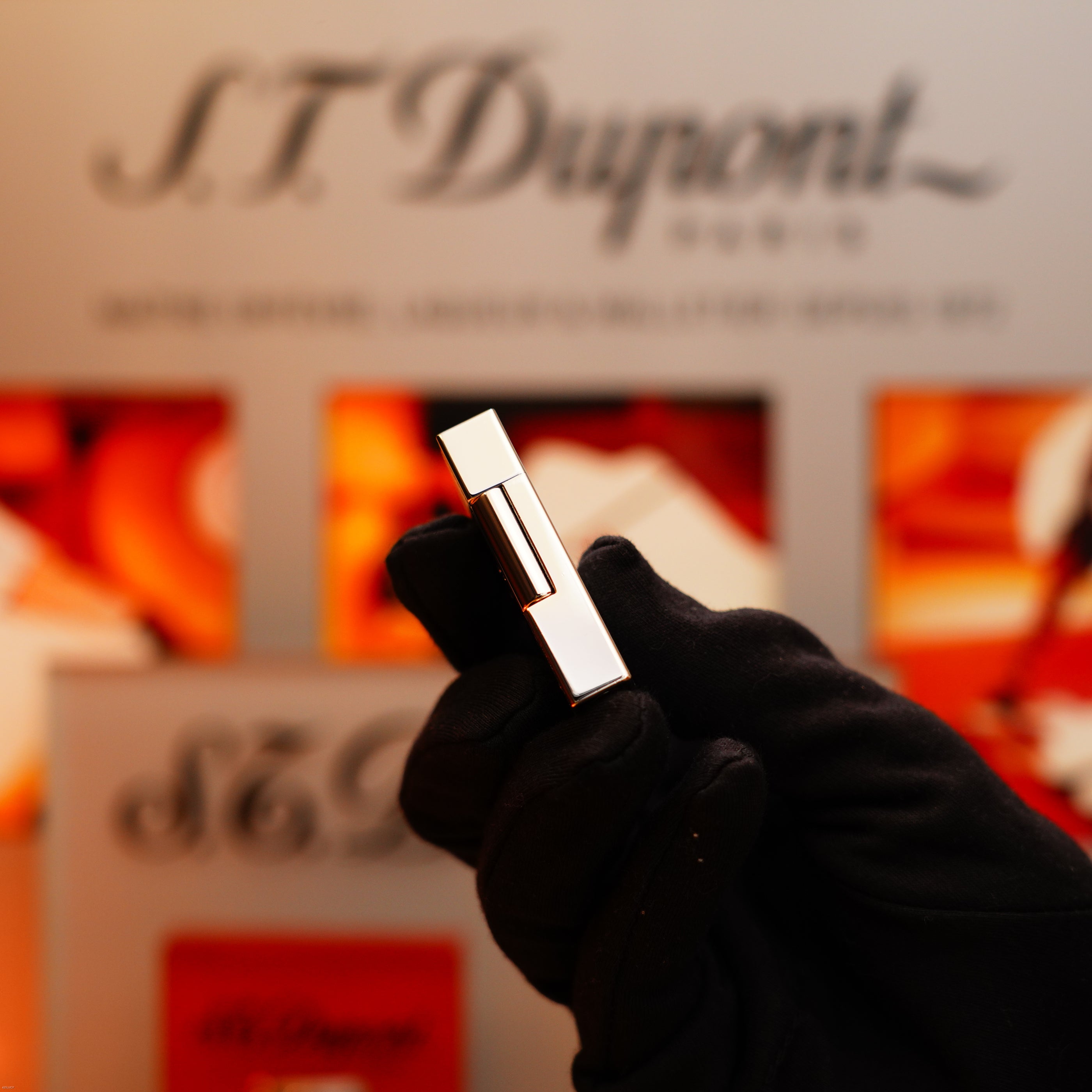 A gloved hand holds a shiny Vintage 2005 S.T. Dupont Gatsby Diamond Drops Limited Edition Lighter, adorned with 16 natural diamonds and heavy platinum, with orange S.T. Dupont packaging blurred in the background.