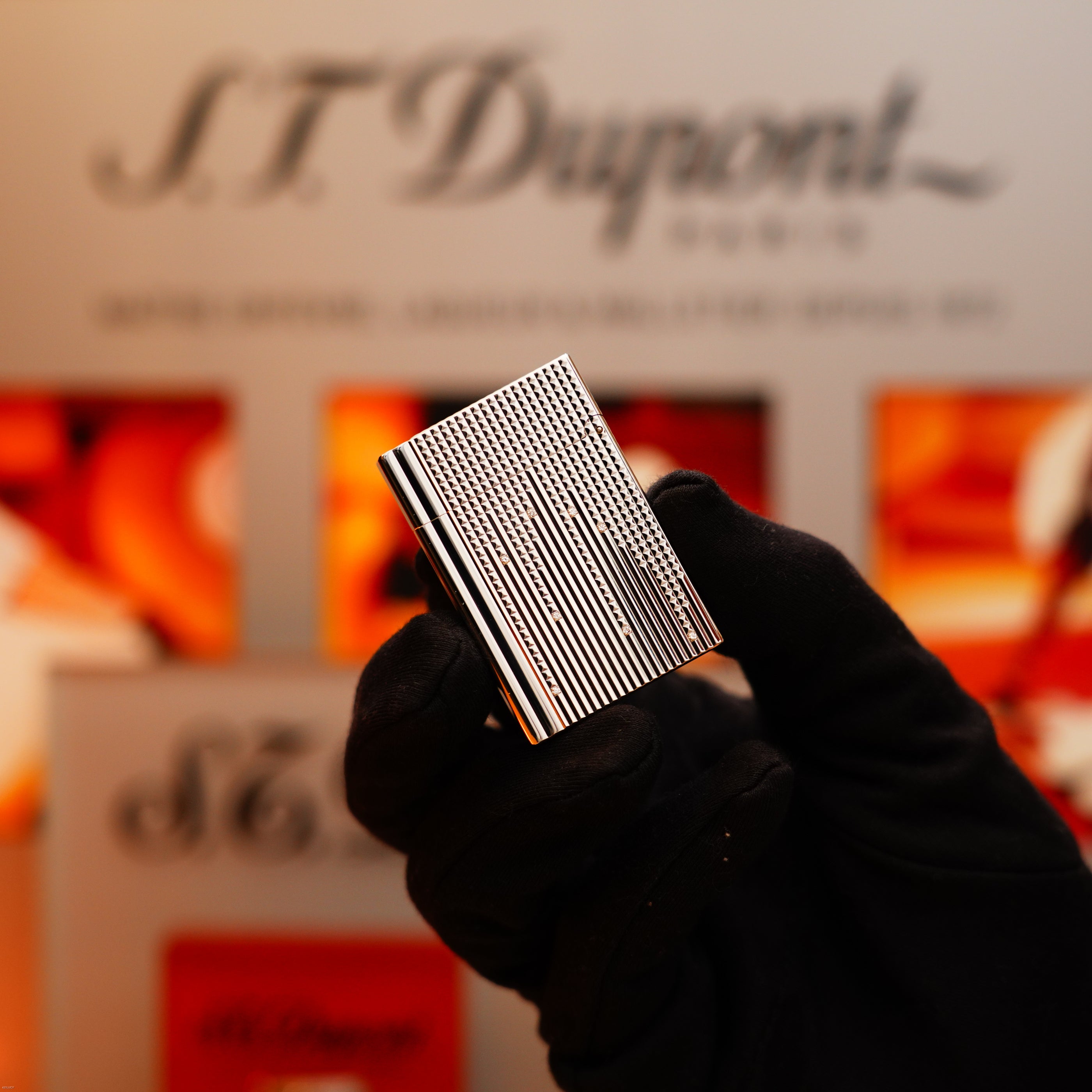 A gloved hand holds the S.T. Dupont Vintage 2005 Gatsby Diamond Drops Platinum Limited Edition Lighter, featuring 16 natural diamonds, in front of a blurred S.T. Dupont sign and red-orange background.