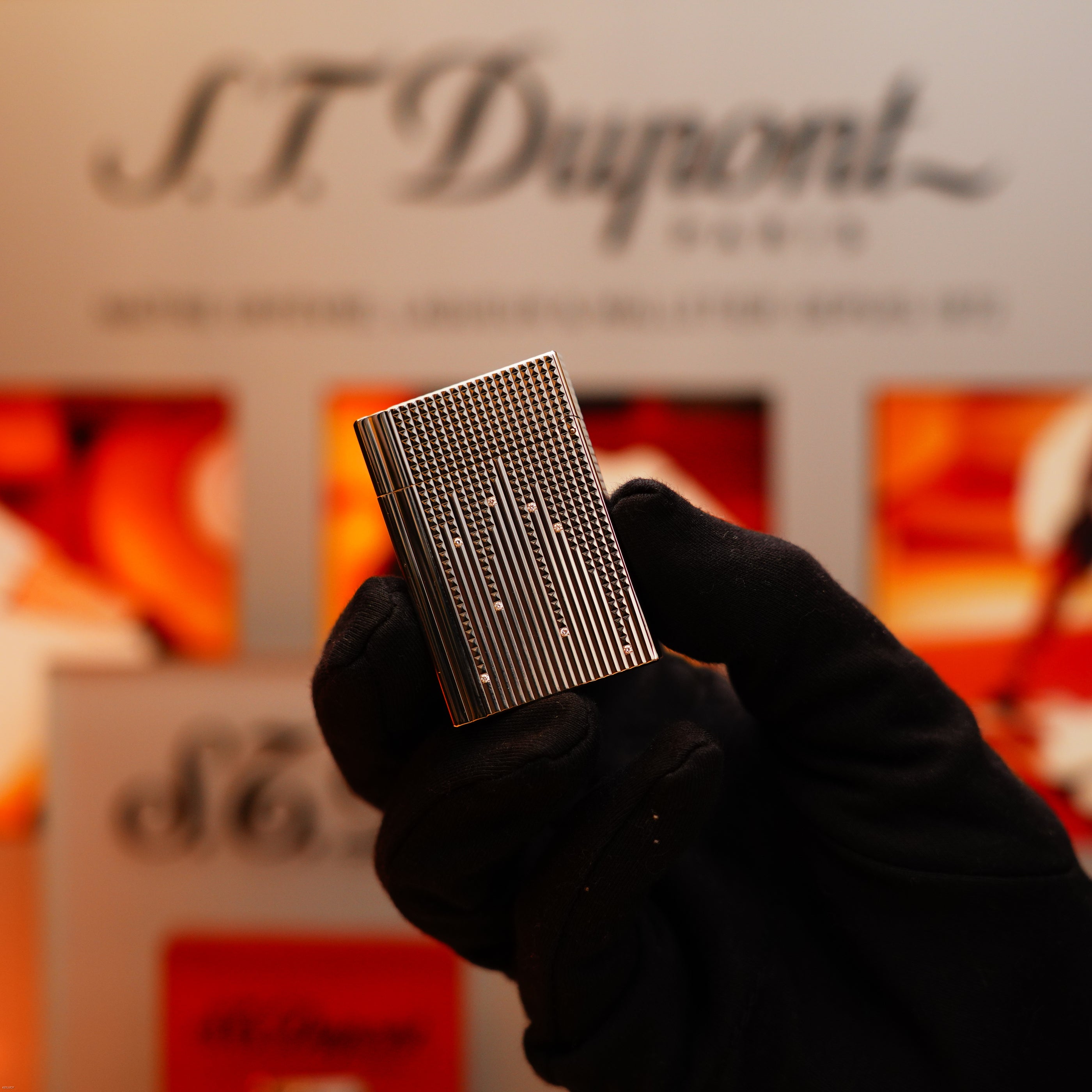 A person wearing black gloves holds a Vintage 2005 S.T. Dupont Gatsby Diamond Drops with 16 Natural Diamond Heavy Platinum Limited Edition Lighter, with a blurred S.T. Dupont sign in the background.
