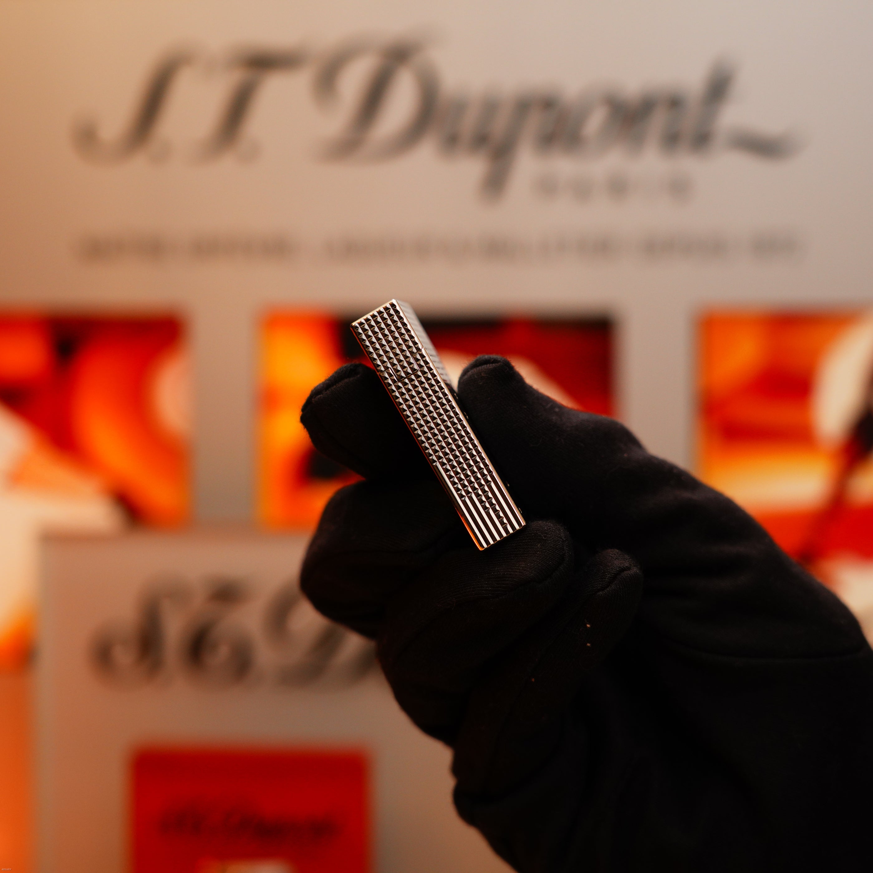 A gloved hand holds the S.T. Dupont Vintage 2005 Gatsby Diamond Drops Limited Edition Lighter, set with 16 natural diamonds and heavy platinum, in front of a blurred S.T. Dupont display background.