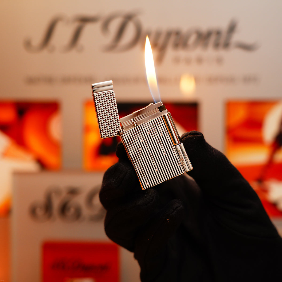 A gloved hand holds a lit S.T. Dupont Vintage 2005 Gatsby Diamond Drops Heavy Platinum Limited Edition Lighter with 16 natural diamonds, in front of a blurred branded display background.